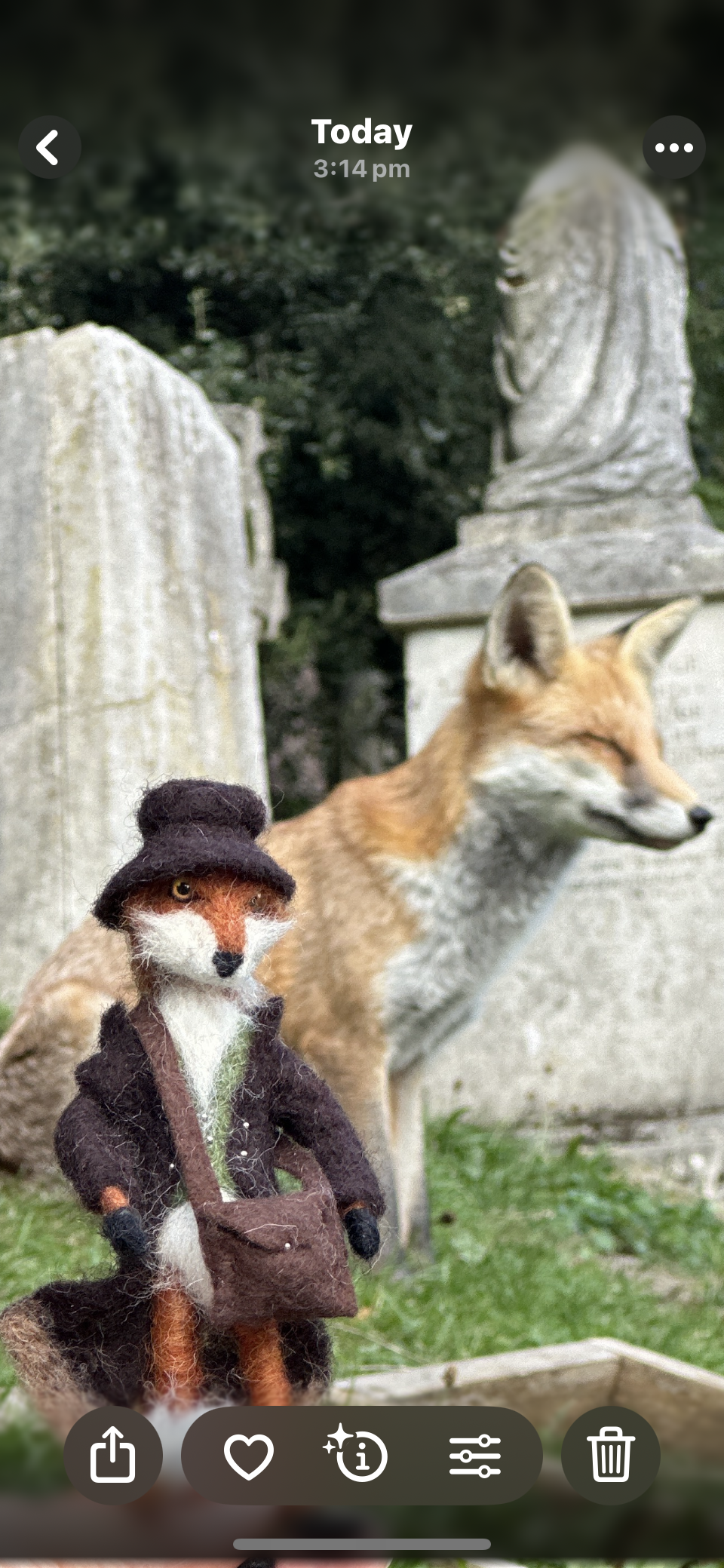 A fox wearing a small black hat and a brown coat with a satchel, standing outdoors in front of stone statues of religious figures, with a blurred background of trees.