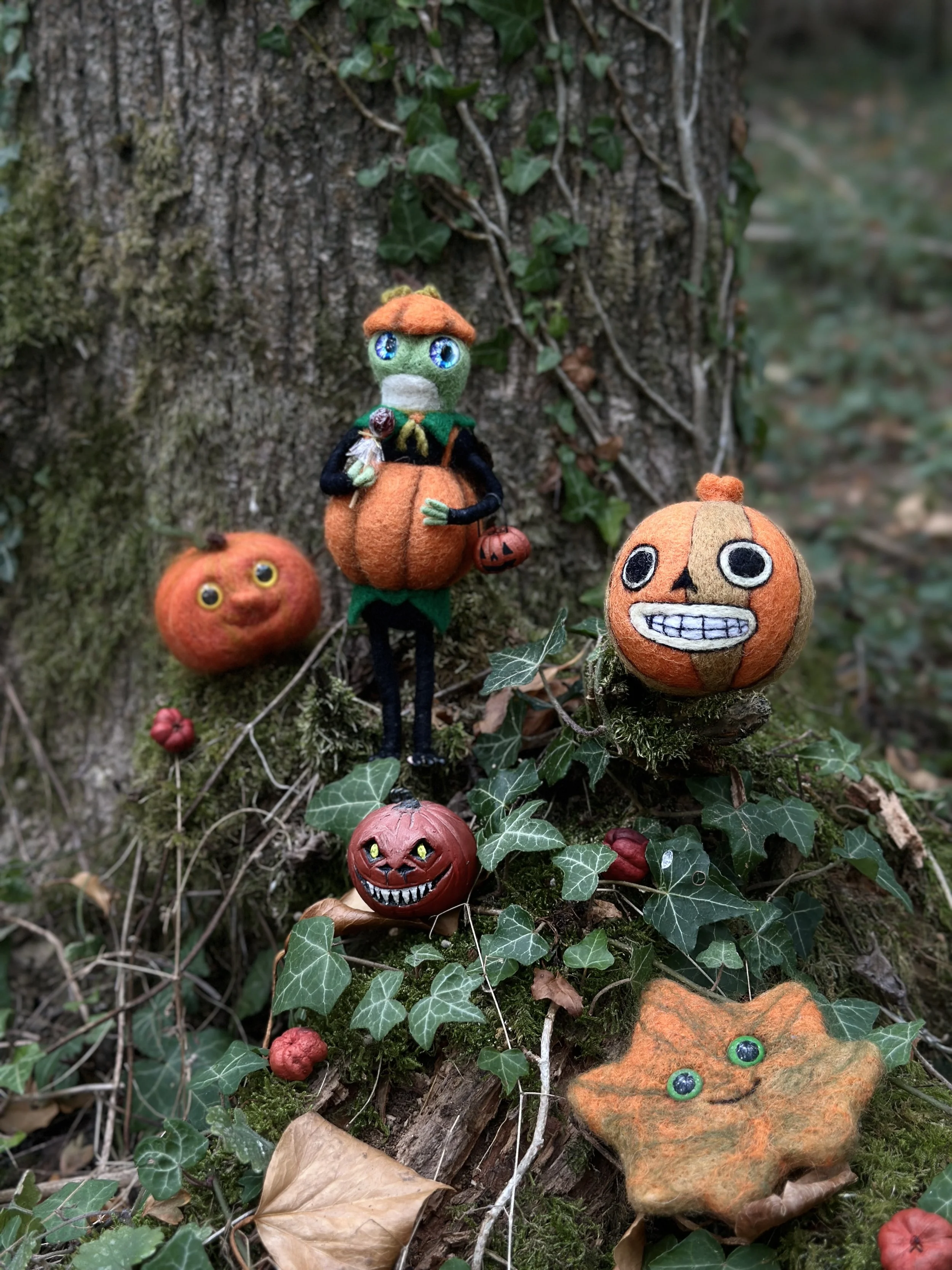 Decorative Halloween-themed felt figures and accessories on a tree trunk, including pumpkin characters with faces, a little witch with bright blue eyes, and autumn leaves, surrounded by green ivy.