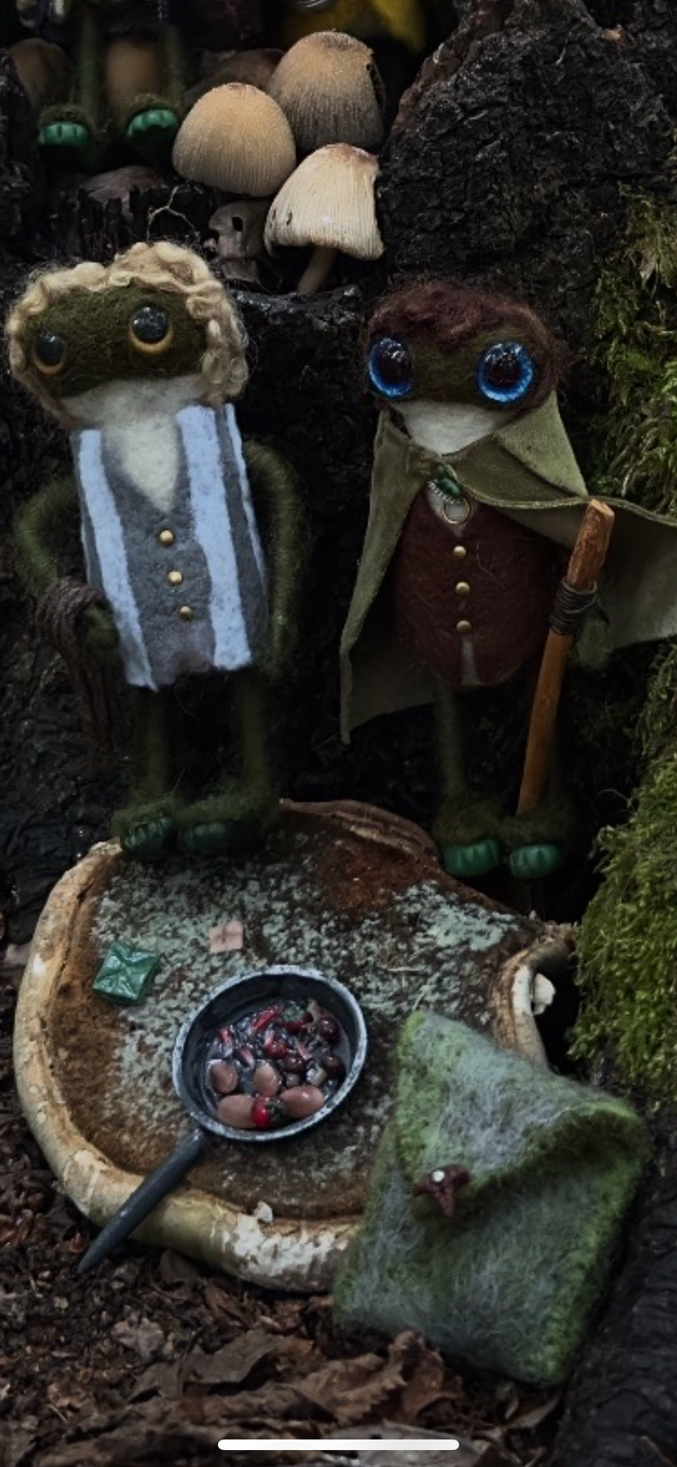 Two plush doll-like figures with large eyes, dressed in fantasy or medieval clothing, standing near a mushroom and a moss-covered rock with a small dish of berries or nuts on a tree stump.