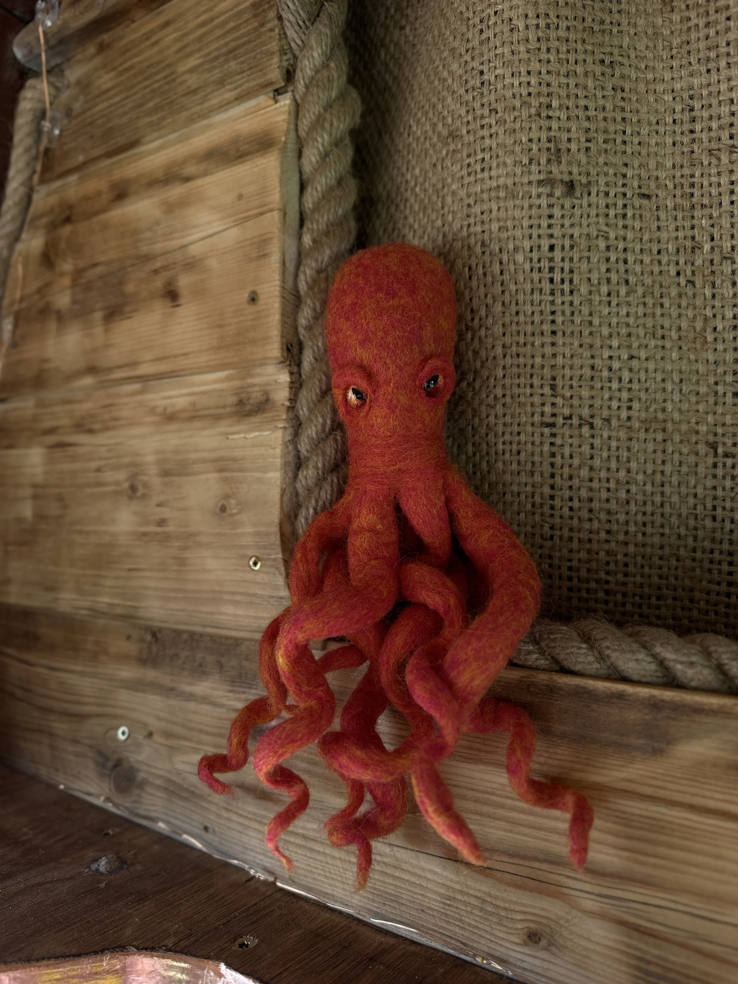 A decorative, felted octopus with red and yellow colors, hanging on a wooden wall with burlap and rope accents.
