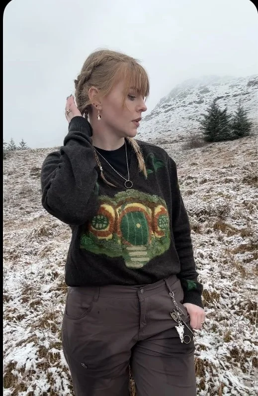 A woman standing outdoors in a snowy landscape with trees and a mountain in the background. She has blonde hair styled in braids and is wearing a black sweater with a colorful, whimsical house design, dark pants, and jewelry.