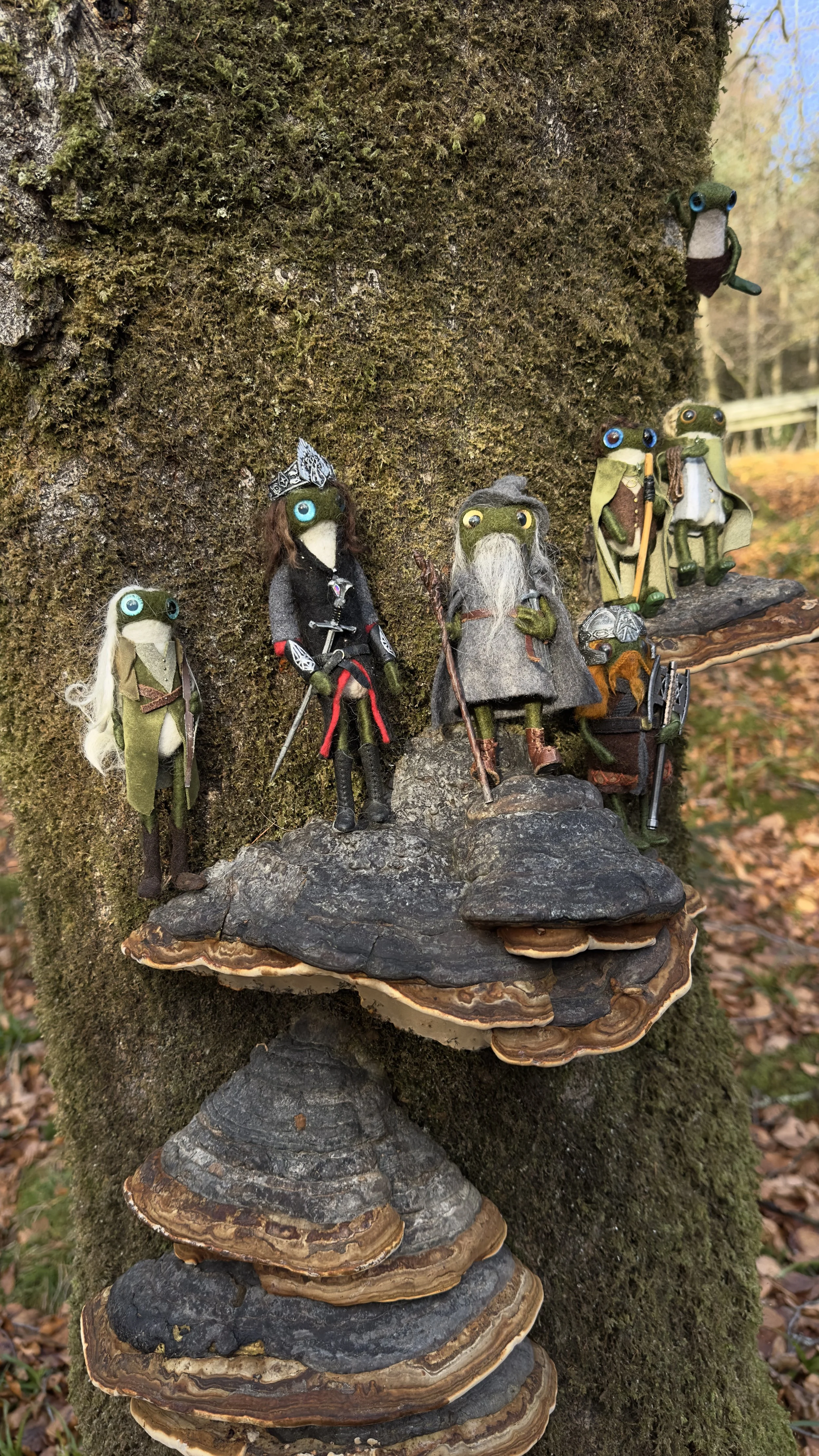 Collection of small handmade frog figurines with distinctive eyes, dressed in various costumes, placed on a tree trunk with mushrooms in the background.