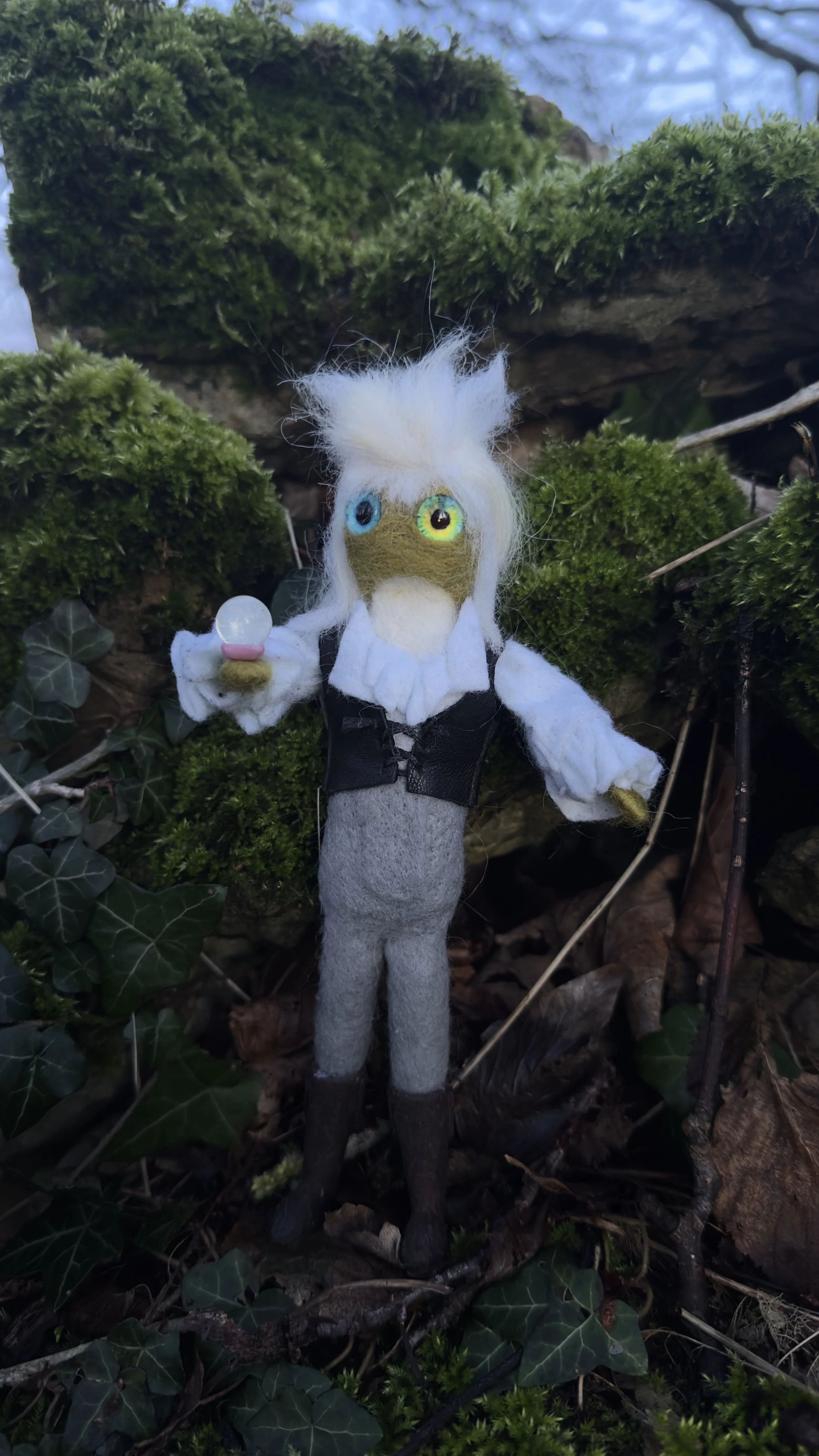 A felted doll resembling a scientist or wizard with wild white hair, colorful eyes, holding a small orb in one hand, standing among moss and ivy on a forest floor.