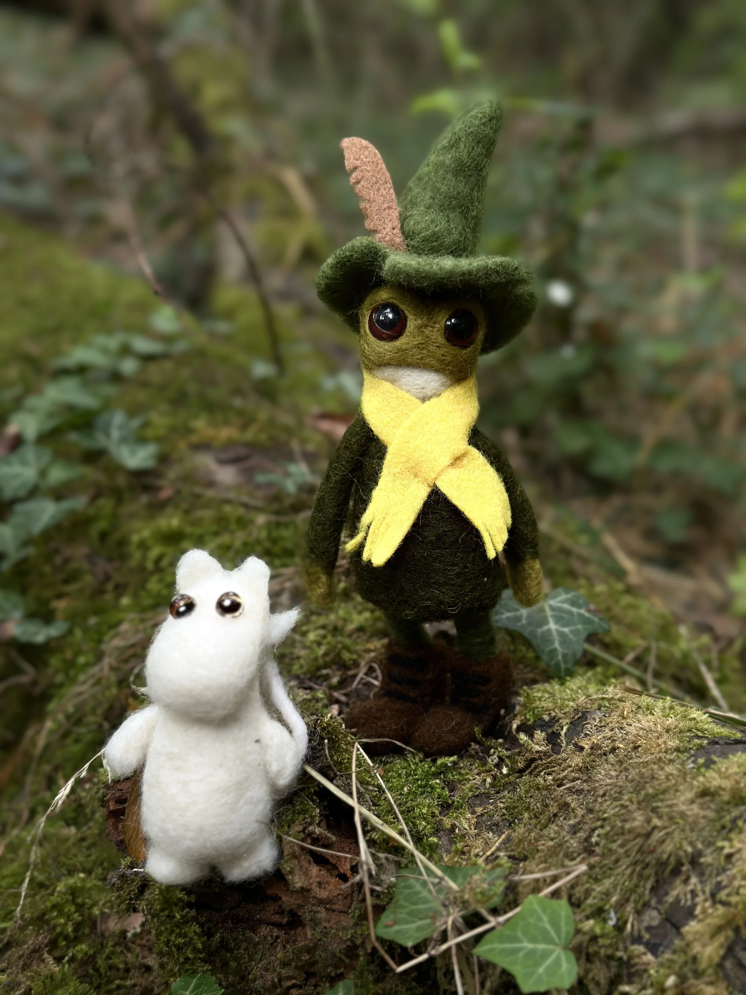 Felted animal figures, including a large green and yellow creature wearing a green hat with a feather and a small white animal, are situated on a mossy log with ivy in a forest setting.