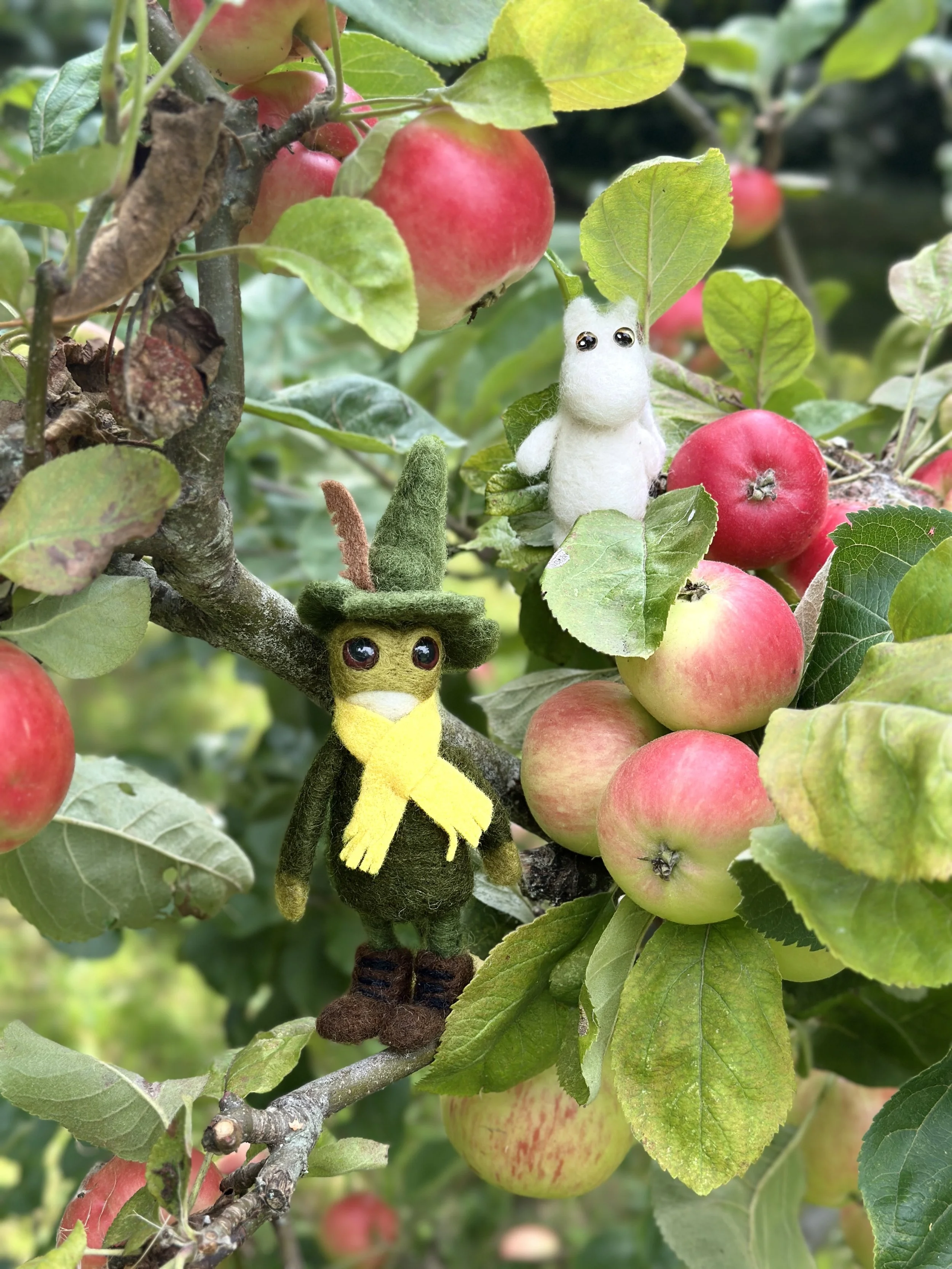 Felted dolls of a white hippo and a green wizard with a hat and scarf, hanging on an apple tree with red and green apples.
