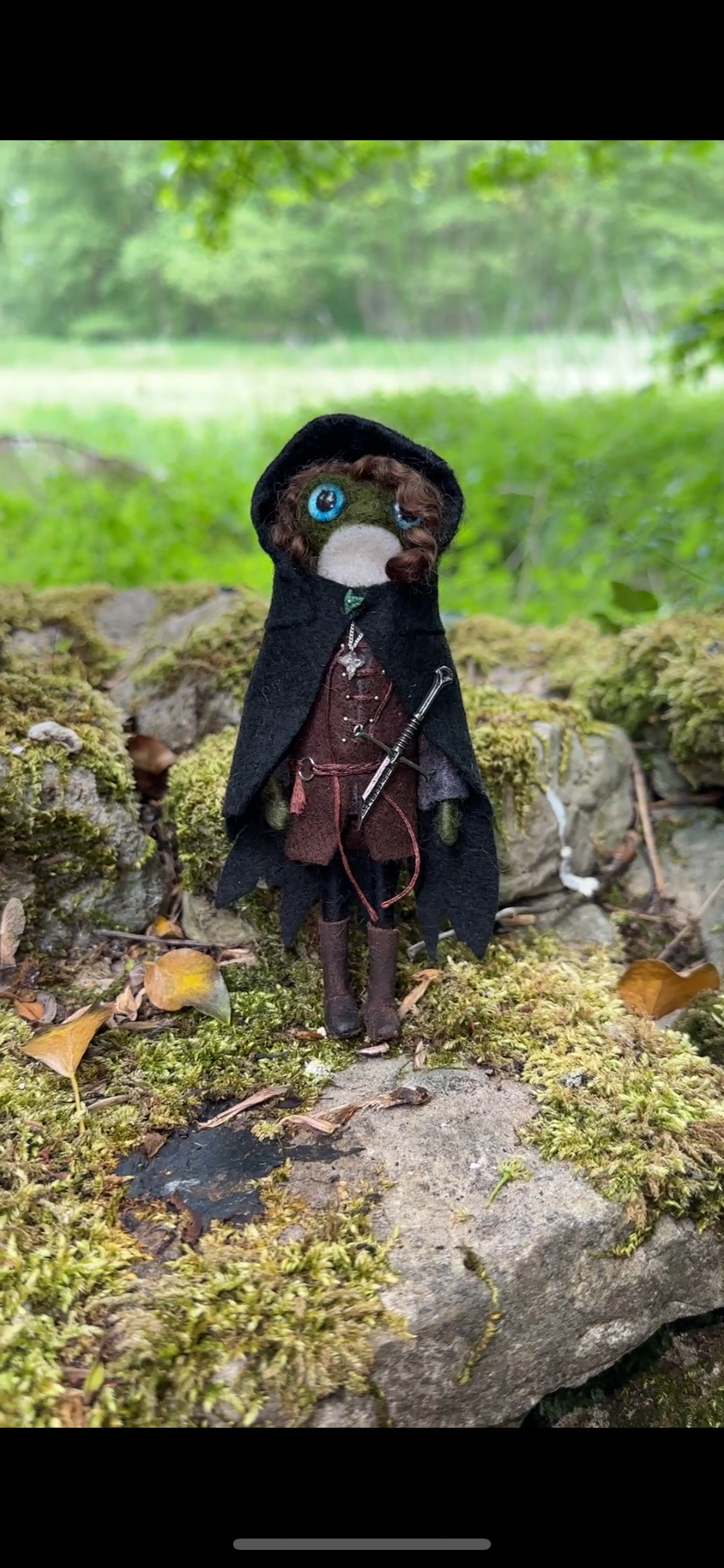 A handmade doll resembling a fantasy character stands outdoors on a moss-covered rock in a green forest. The doll has blue eyes, brown curly hair, and wears a dark cloak, brown outfit, and is equipped with a small sword.