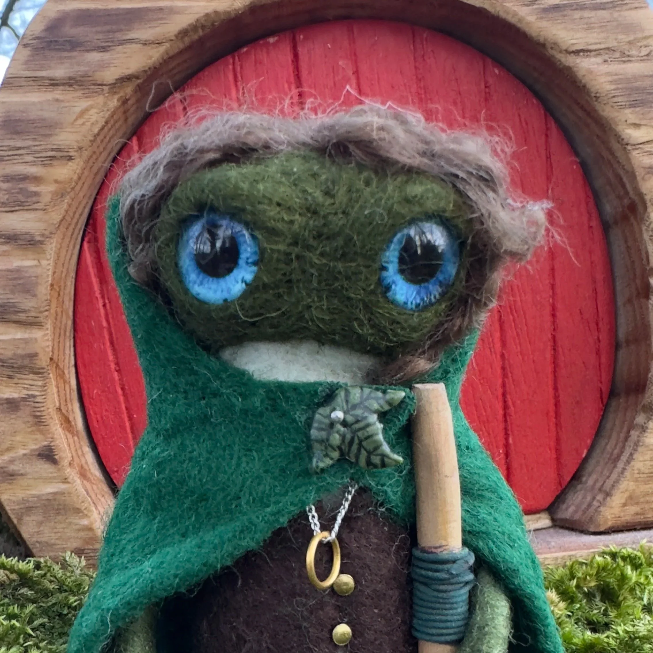 Felted doll resembling an elf or fantasy character with large blue eyes, wearing a green cloak, holding a staff, and standing in front of a red circular wooden background.