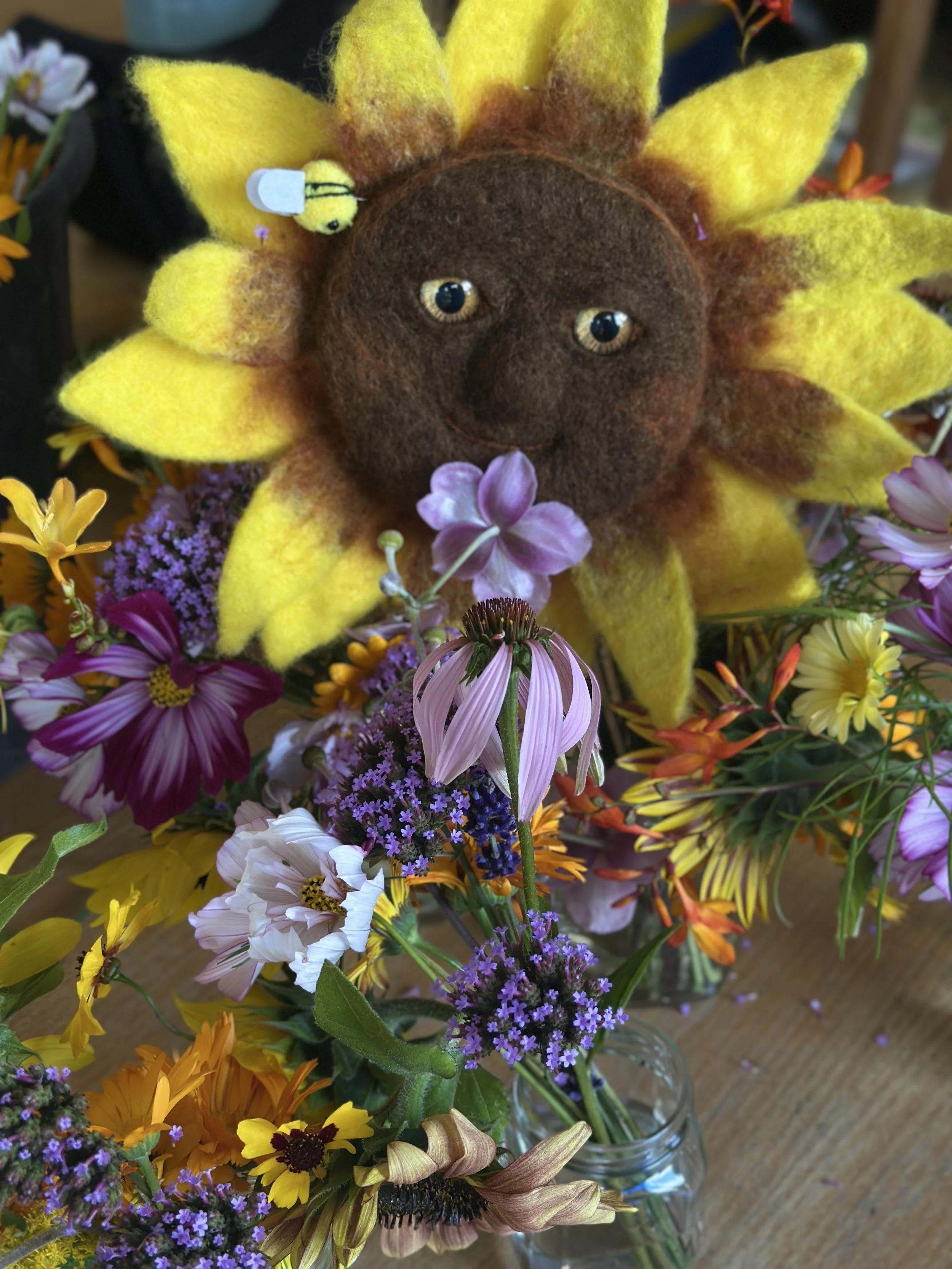 Felt sunflower with a smiling face, surrounded by various colorful flowers in a vase.