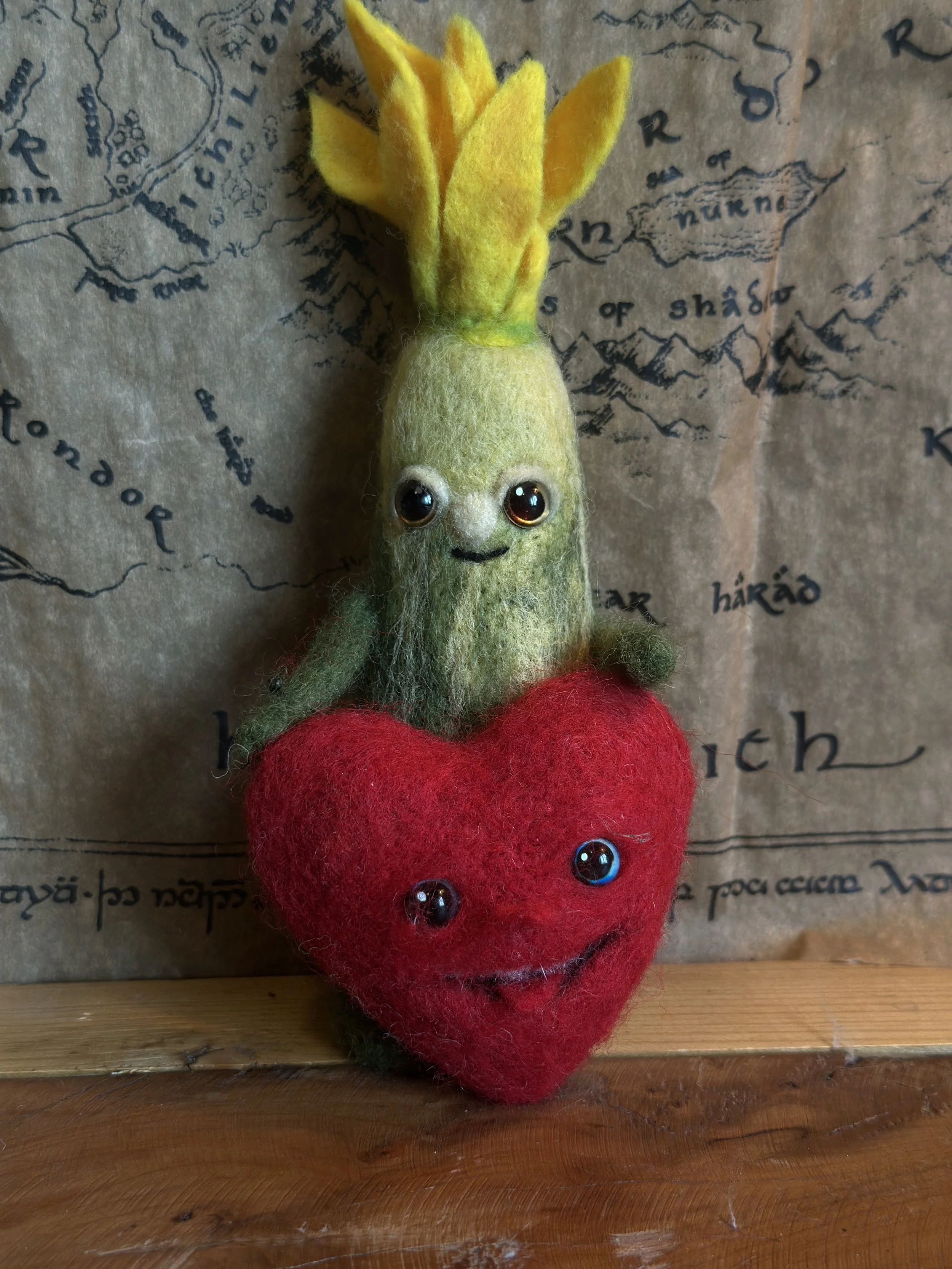 Felted plush toy in the shape of a cheerful zucchini with a yellow flower top, sitting on a red heart with a smiling face, set against a vintage map background.