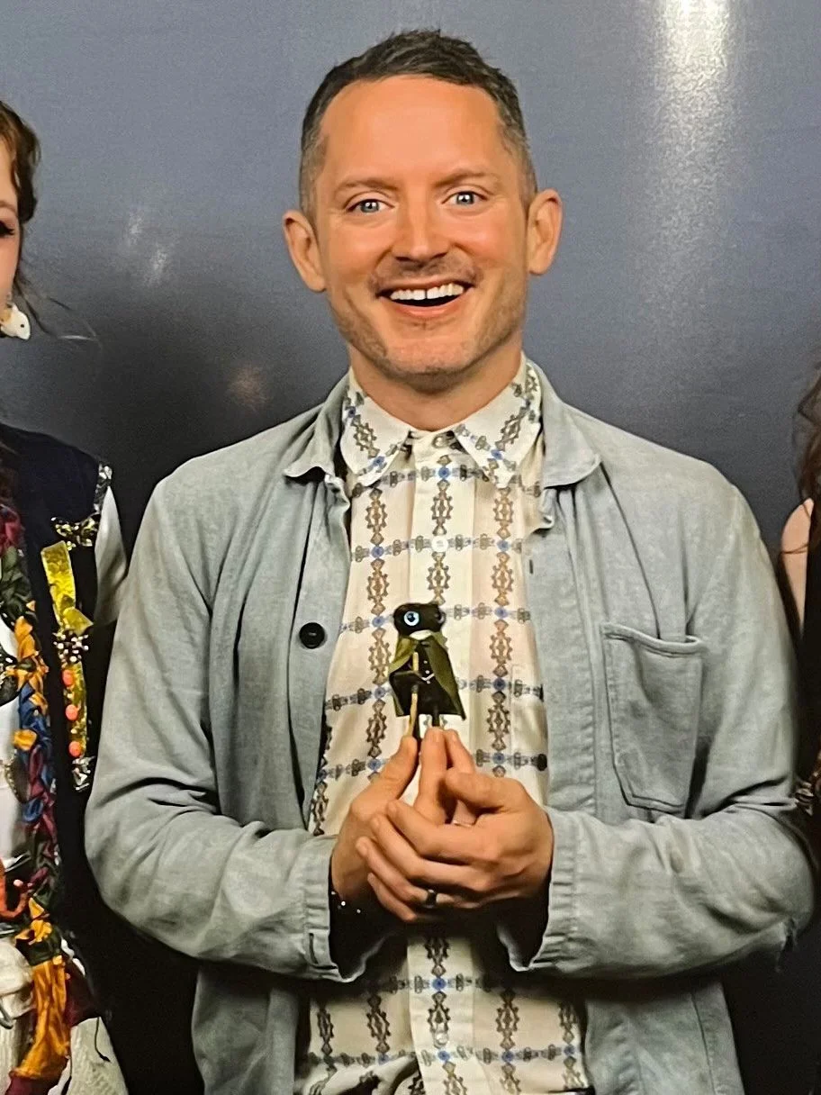A smiling man with short hair holding a small figurine or doll, standing against a dark background.