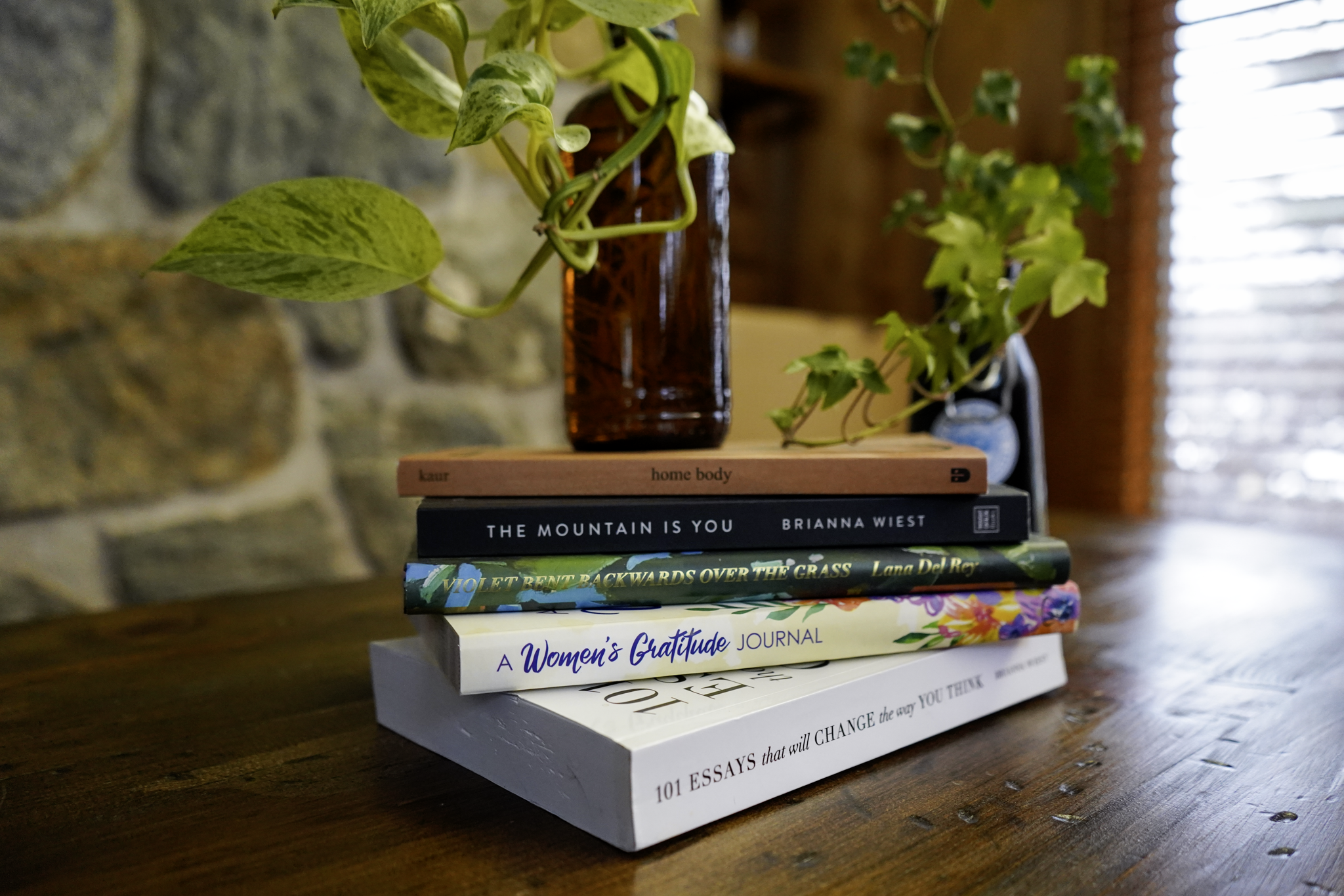 Stack of five books on a wooden table with a brown glass vase containing green and variegated leaves in the background.