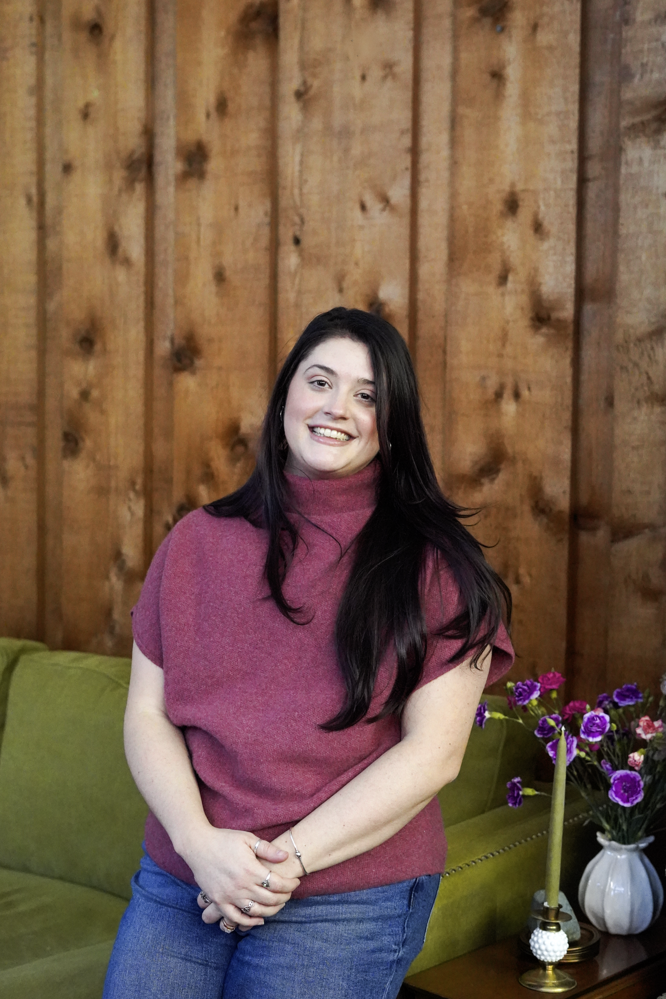 A smiling woman with long dark hair wearing a pink turtleneck sweater and blue jeans, standing indoors in front of a wooden wall, with a green sofa and a vase of purple flowers in the background.