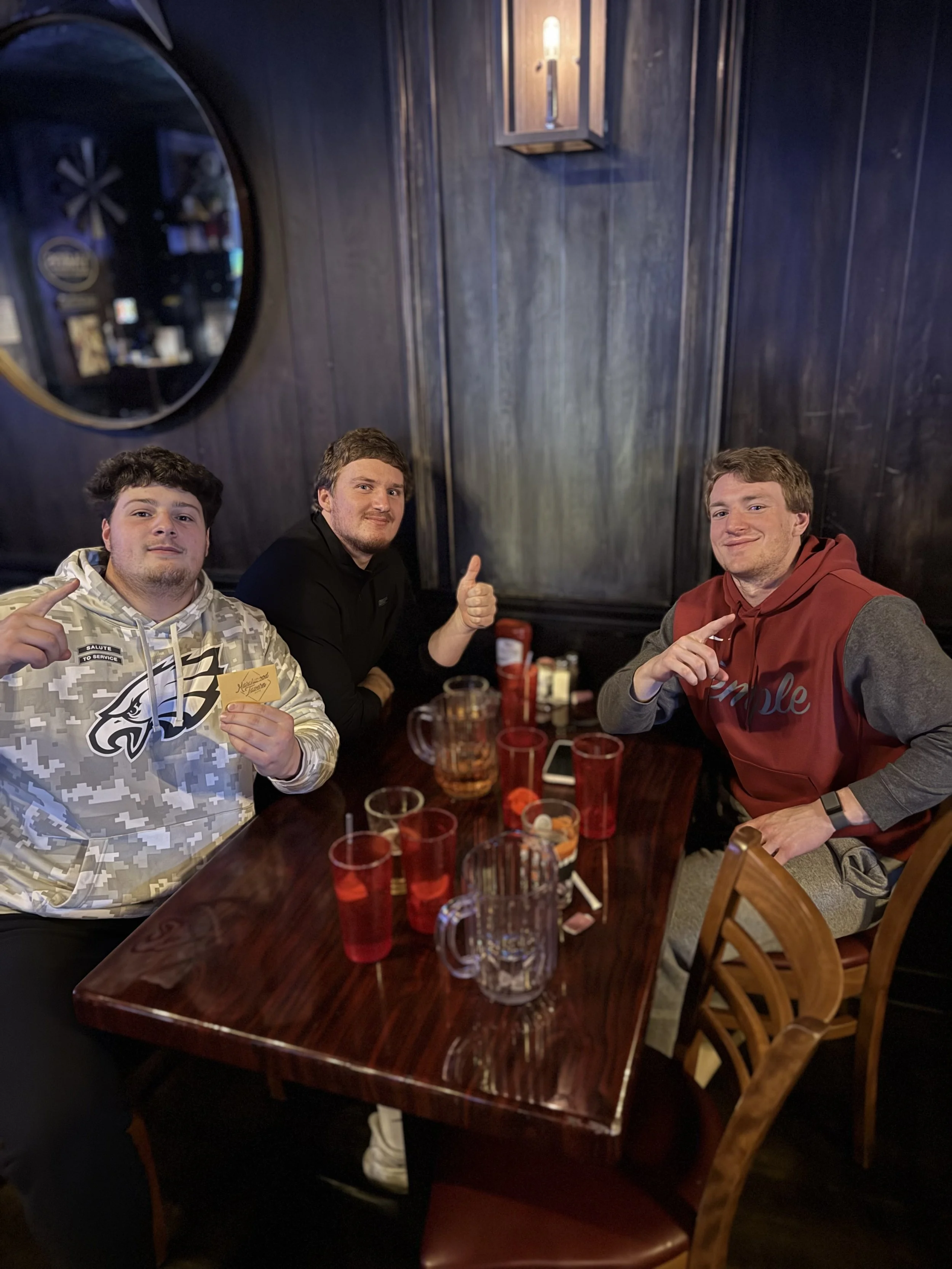 Three young men sitting at a dark wooden table in a dimly lit restaurant or bar, with red cups, pitchers, and a smartphone on the table, giving thumbs up and smiling.