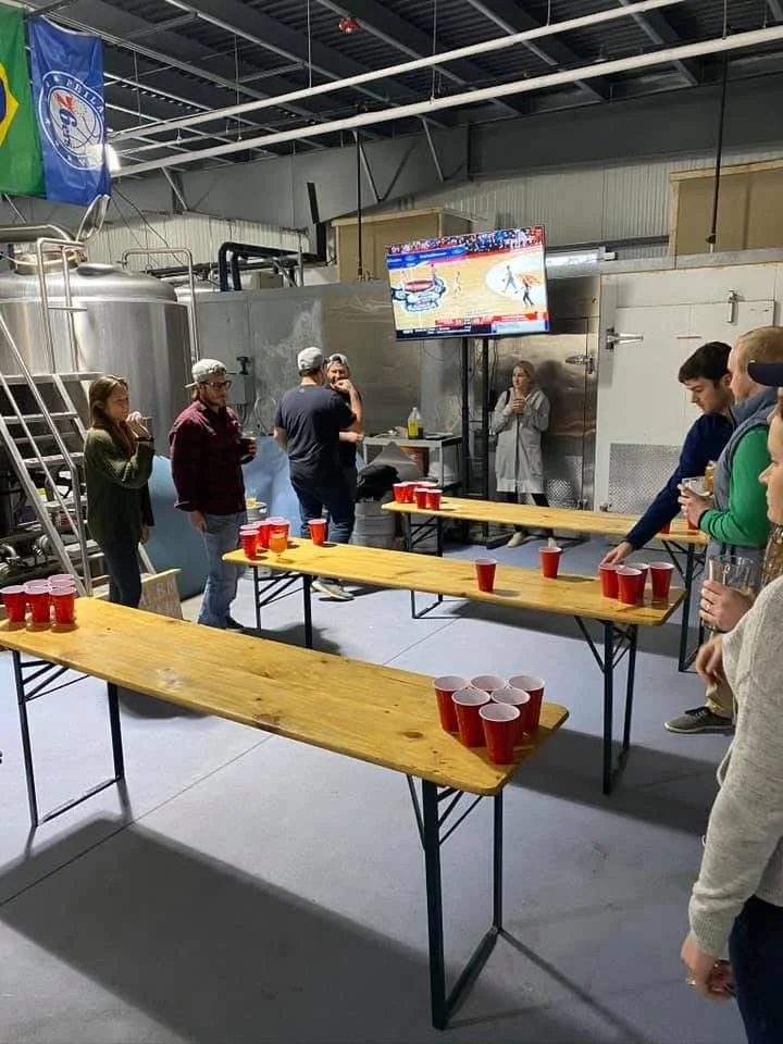 People playing beer pong in an industrial brewery with brewing tanks and a television displaying a hockey game.