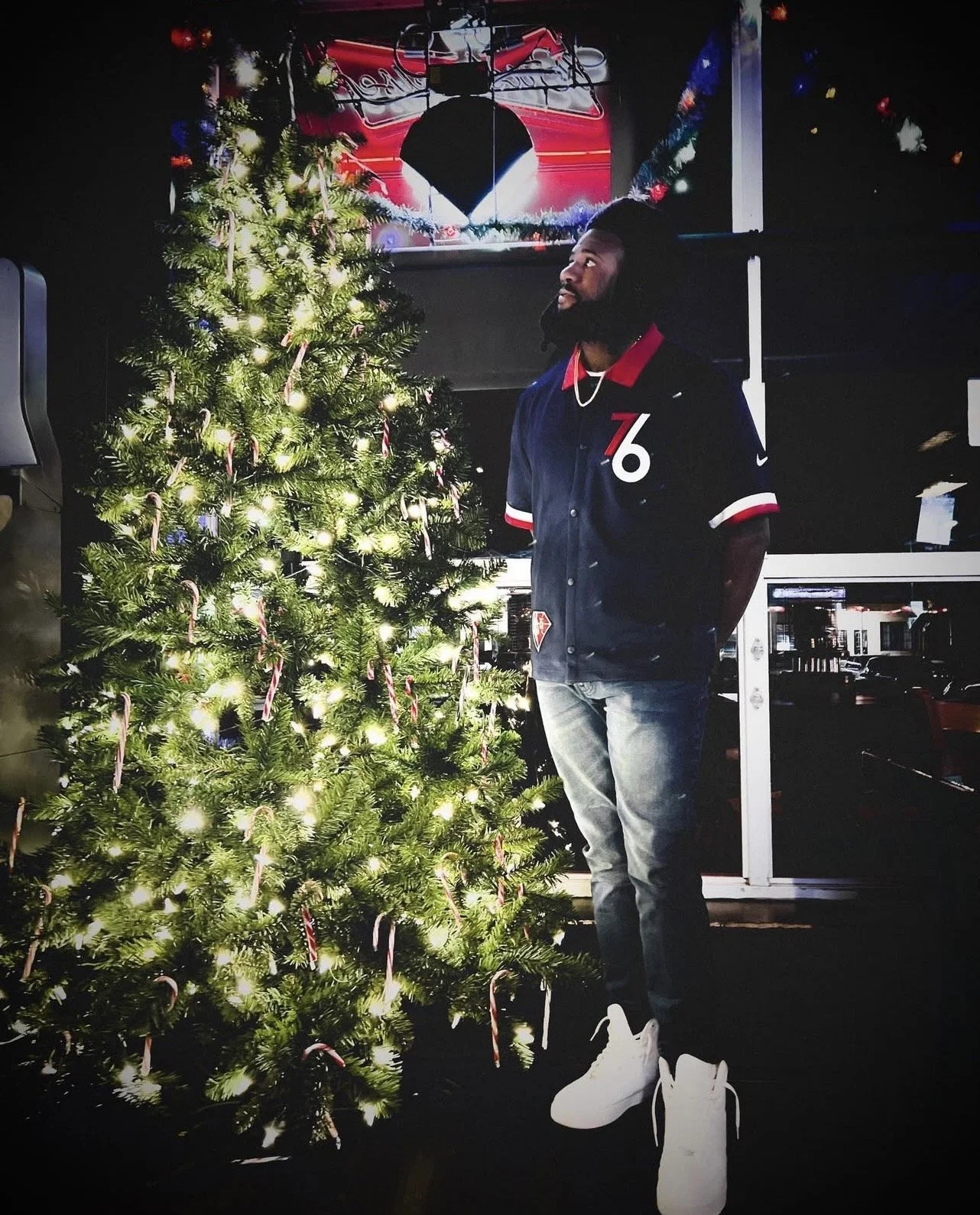 A man standing next to a decorated Christmas tree with candy canes, in a dimly lit room with a neon sign and colorful decorations in the background.