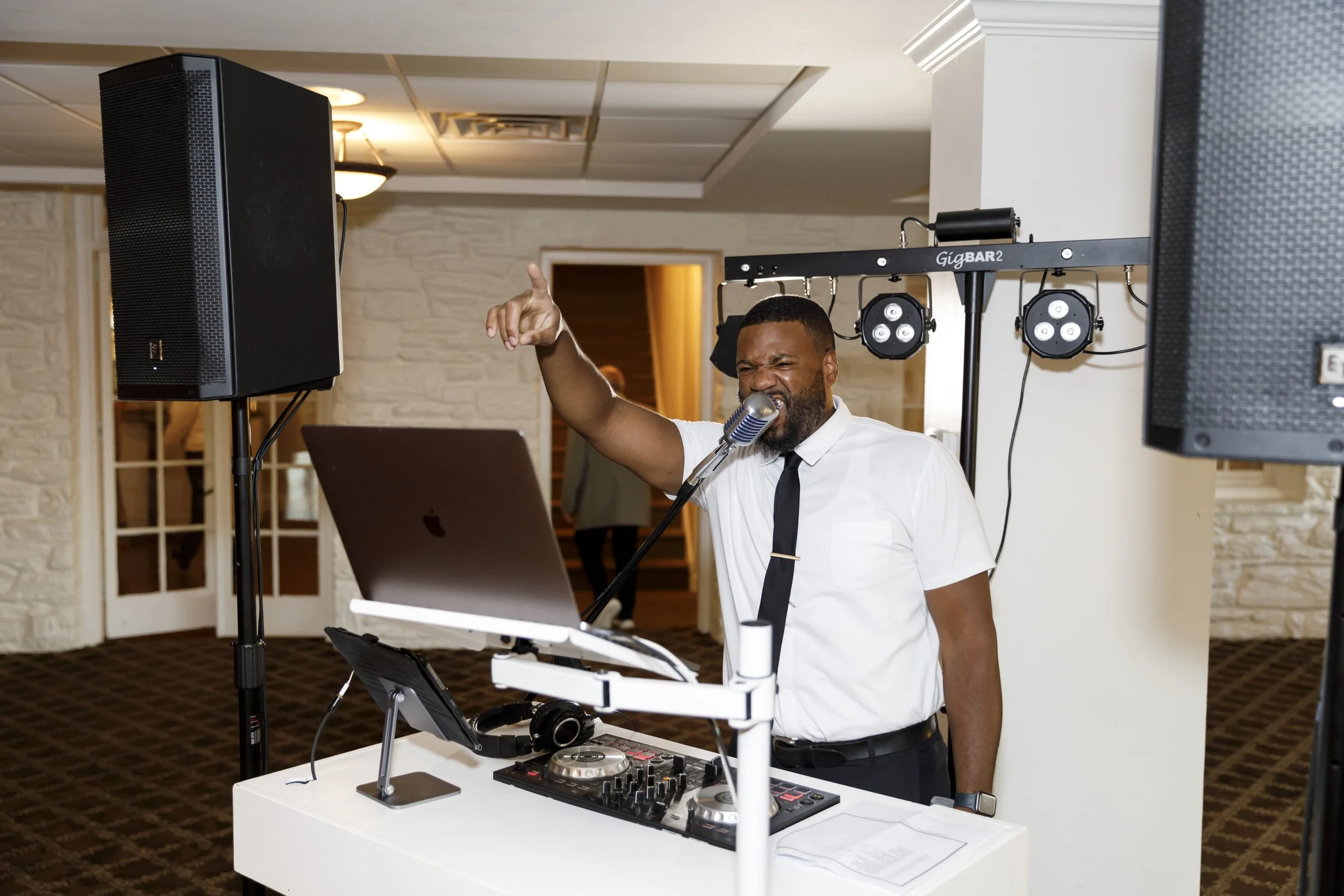 A DJ in a white shirt and black tie is singing into a microphone while mixing music on a DJ controller, with speakers and stage lights around him at an indoor event.