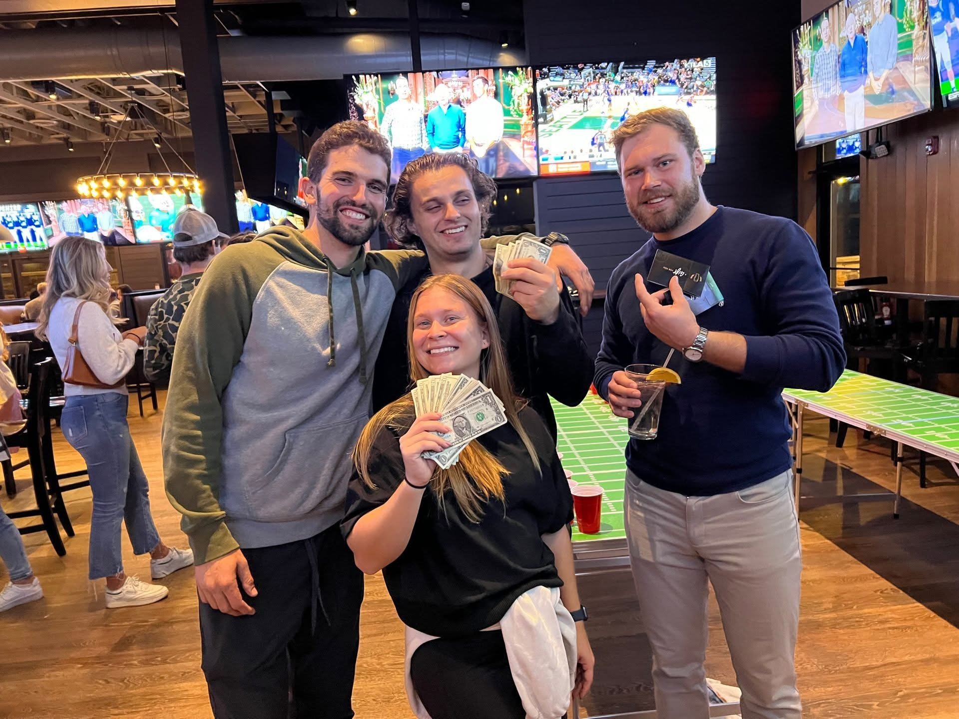 Group of four friends celebrating at a bar, holding cash and drinks, smiling, with TV screens showing sports in the background.