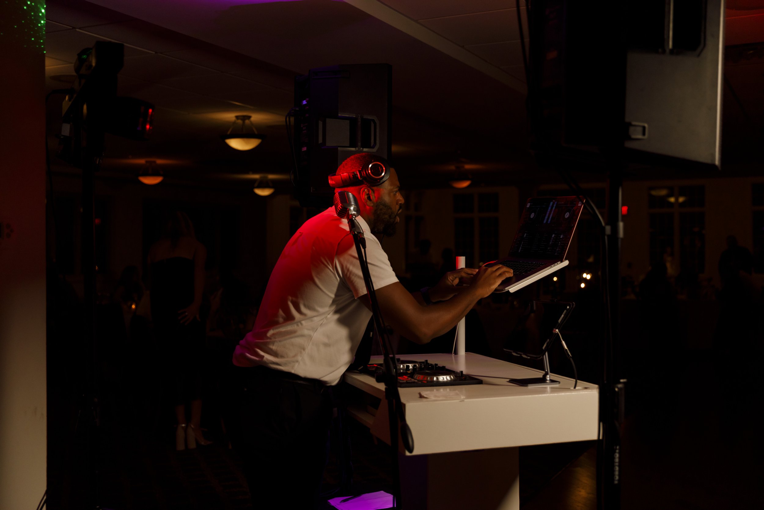 DJ playing music on a laptop at a dimly lit event, with headphones on, in a dark indoor venue.
