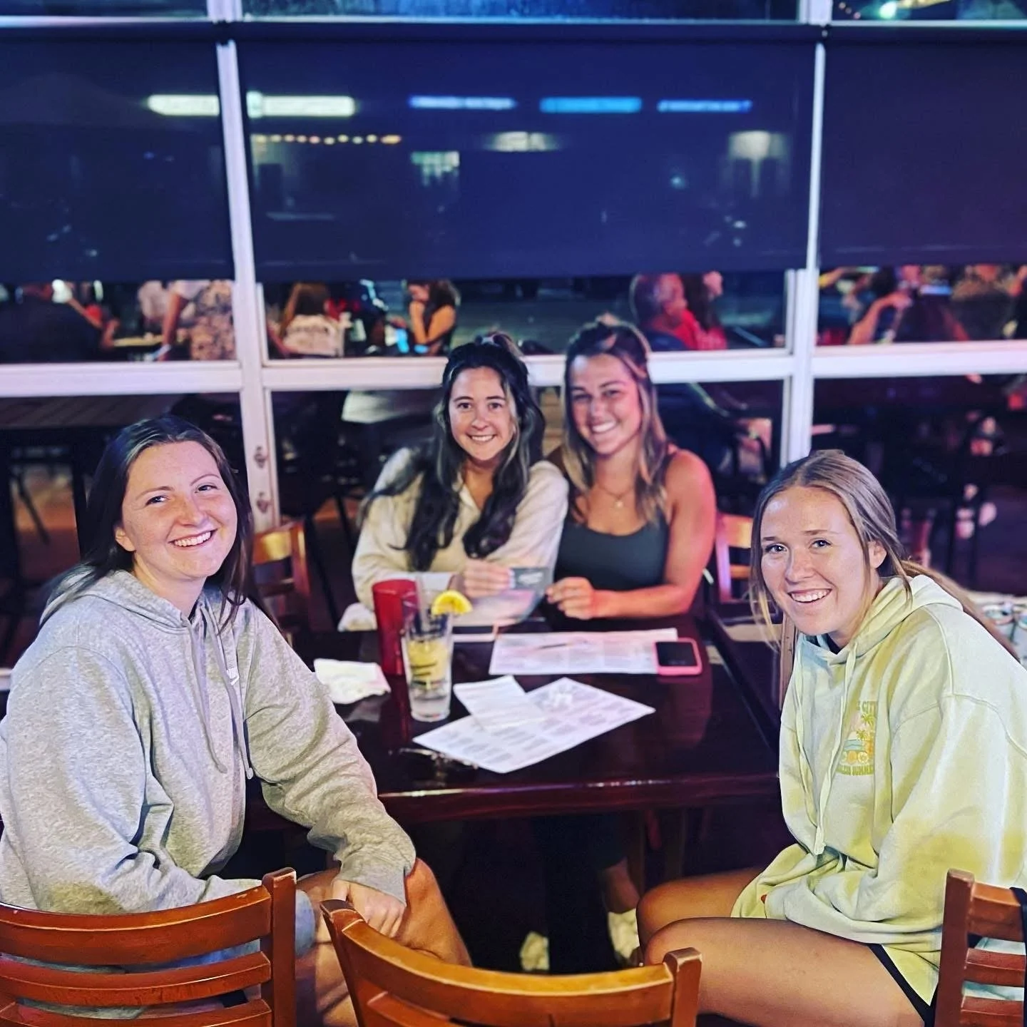 Four young women sitting at a restaurant table, smiling at the camera, with drinks and menus in front of them, in a brightly lit indoor setting with other patrons and a large window behind them.