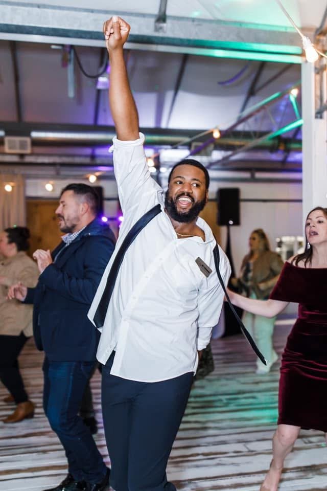 Man with a beard and white shirt dancing with one arm raised at a party or social gathering.