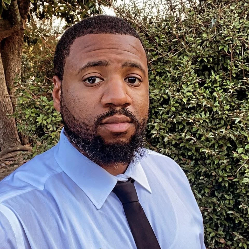 A man with a beard wearing a light blue dress shirt and black tie taking a selfie outdoors in front of green foliage.