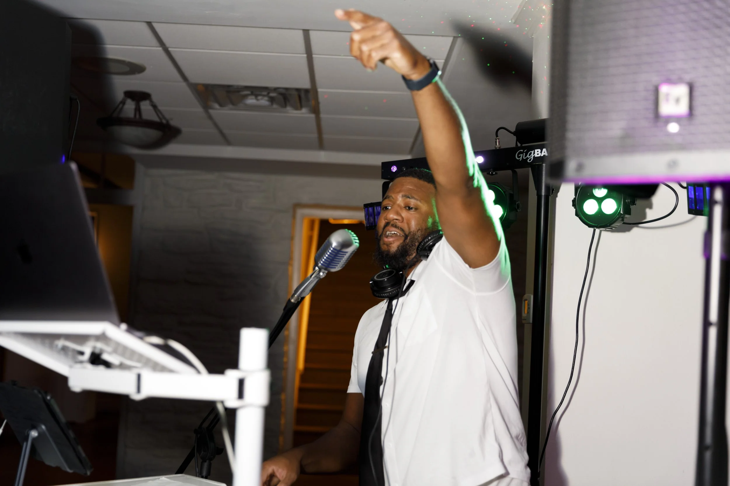 A male DJ wearing a white t-shirt, with headphones around his neck, is performing at a DJ booth. He has a microphone in front of him and is raising one arm while singing or speaking. The scene is lit with green stage lights.