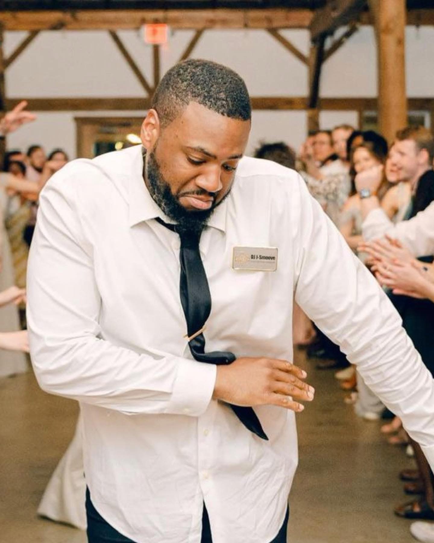 A waiter in a white shirt and black tie, wearing a name tag, appears to be cleaning a table in a lively restaurant or event venue with people sitting at tables in the background.