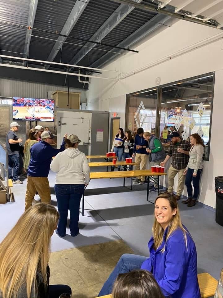 People playing, watching a bean bag tossing game at a social gathering inside a modern industrial-style space with a high ceiling, a large window, and a TV screen.