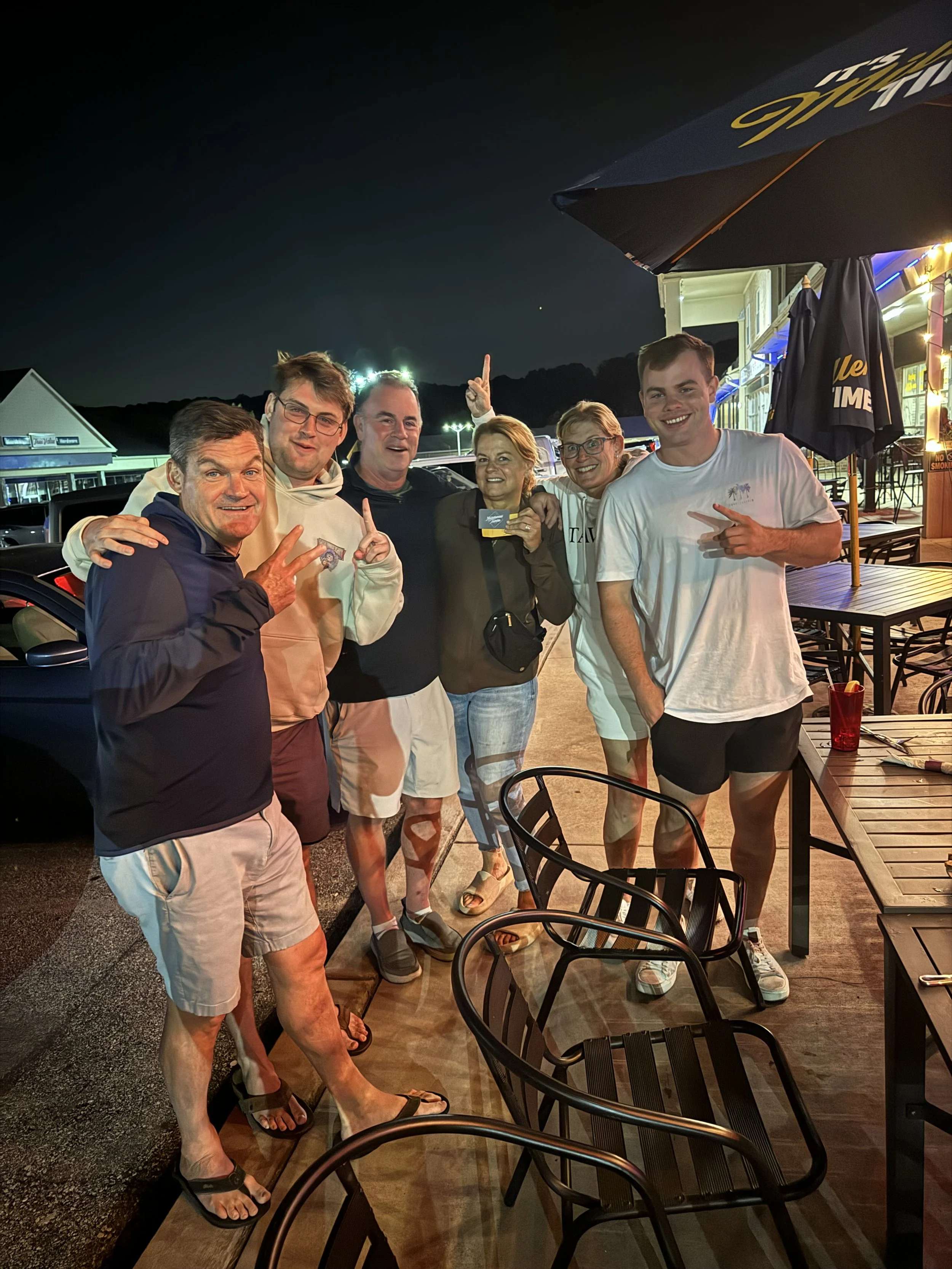 Group of six people standing outside at night, smiling and making gestures, near a table with chairs and a large umbrella, with a parking lot and buildings in the background.