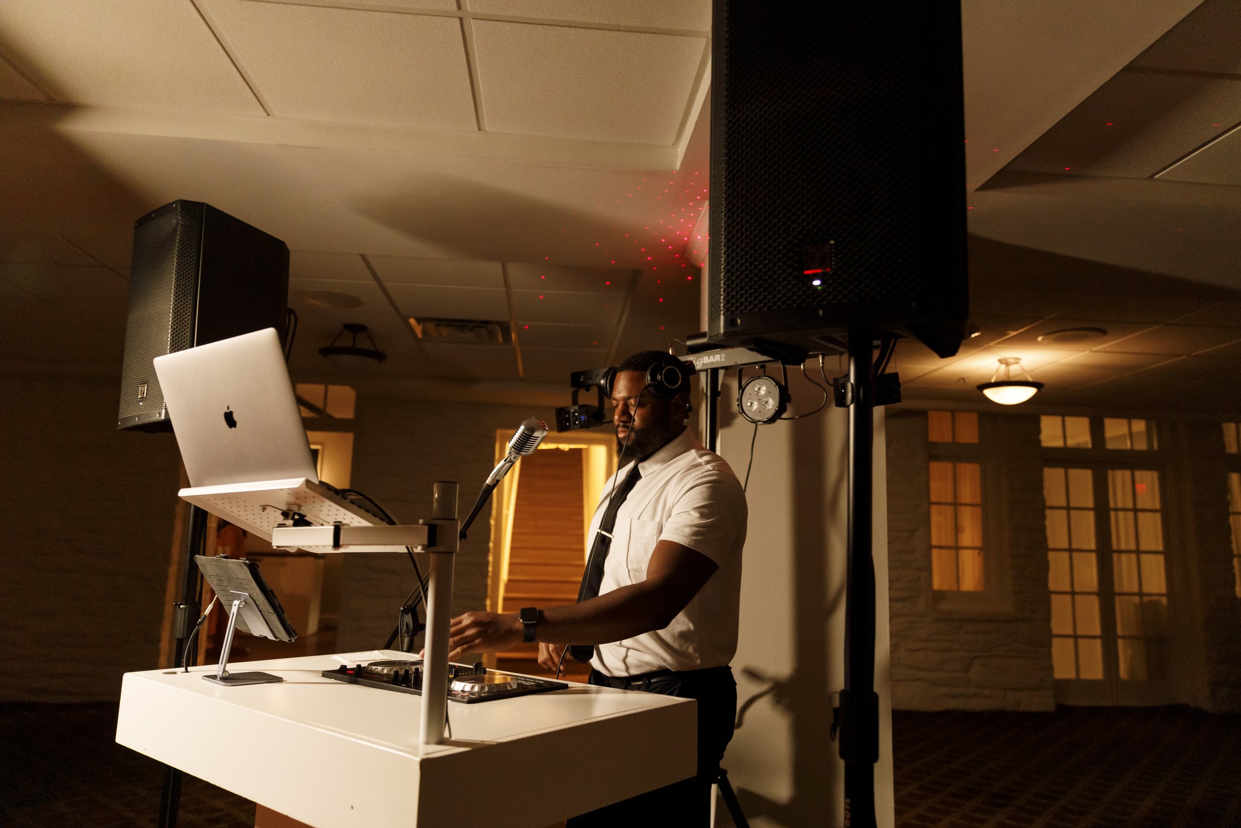 A DJ wearing headphones, a white shirt, and a black tie stands at a DJ booth with a laptop, mixer, and music controls, performing at a dimly lit event space with orange lighting and large windows in the background.