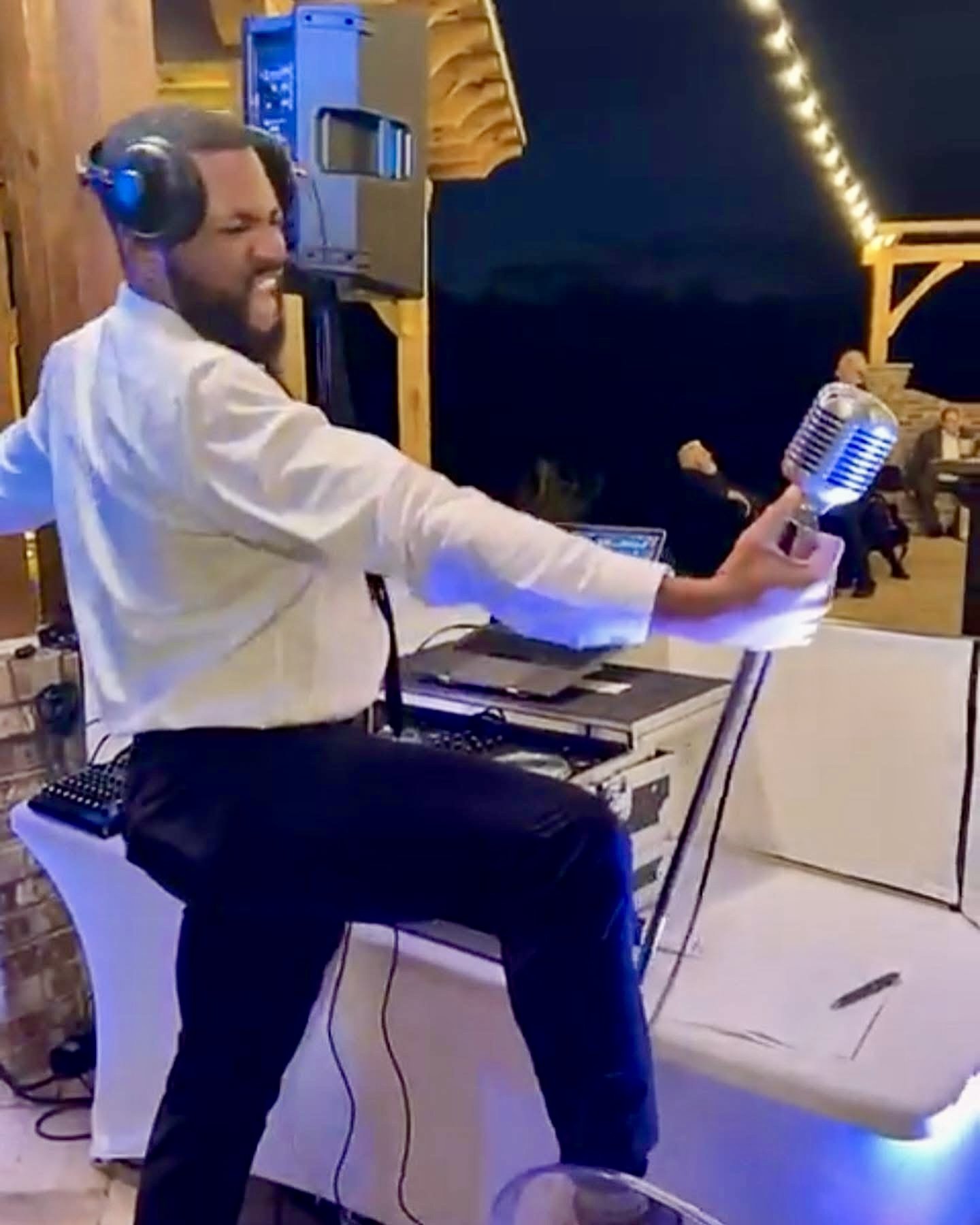 A man in a white shirt and black pants is holding a vintage microphone while wearing large headphones, standing in a dimly lit outdoor venue with string lights in the background.