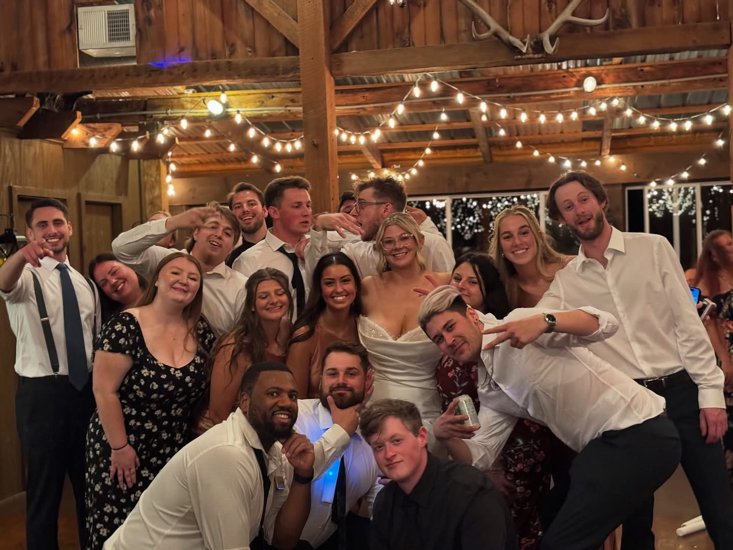 Group of friends celebrating at a party in a rustic venue with string lights, some smiling, some making playful gestures, and a woman in a white dress at the center.