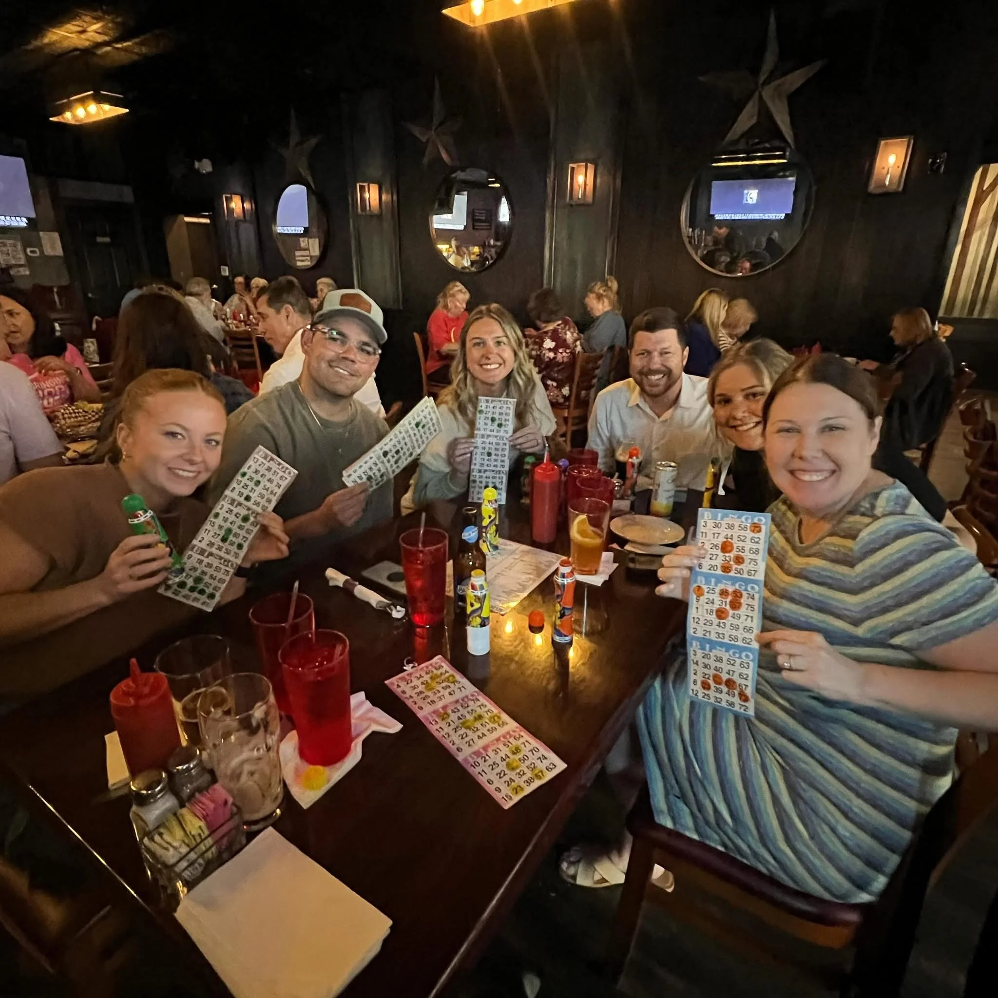 A group of seven people seated around a table in a restaurant, holding bingo cards and smiling for the camera during a bingo night.