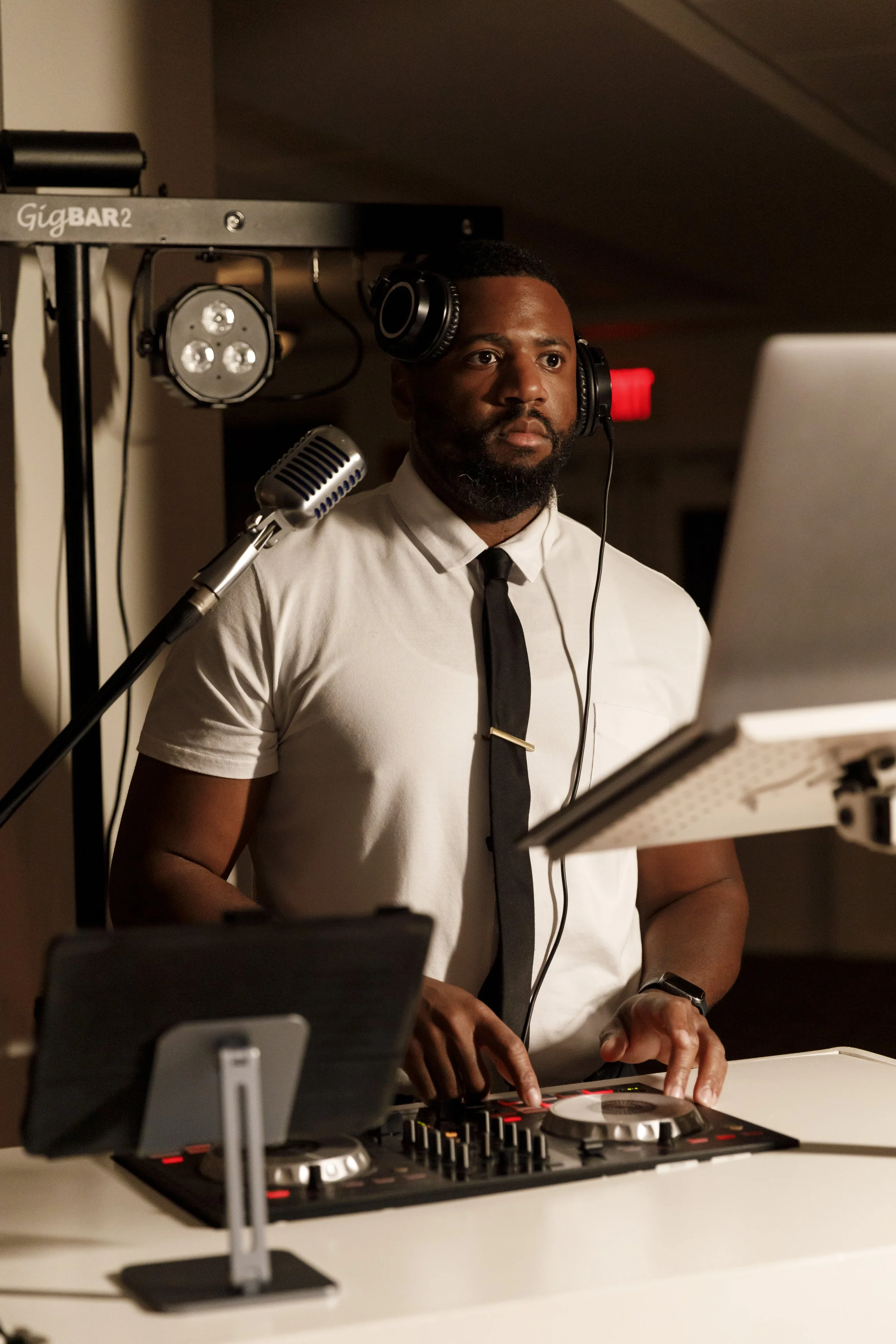 A man with a beard and headphones, standing behind DJ equipment, wearing a white shirt and black tie, in a dimly lit indoor setting.