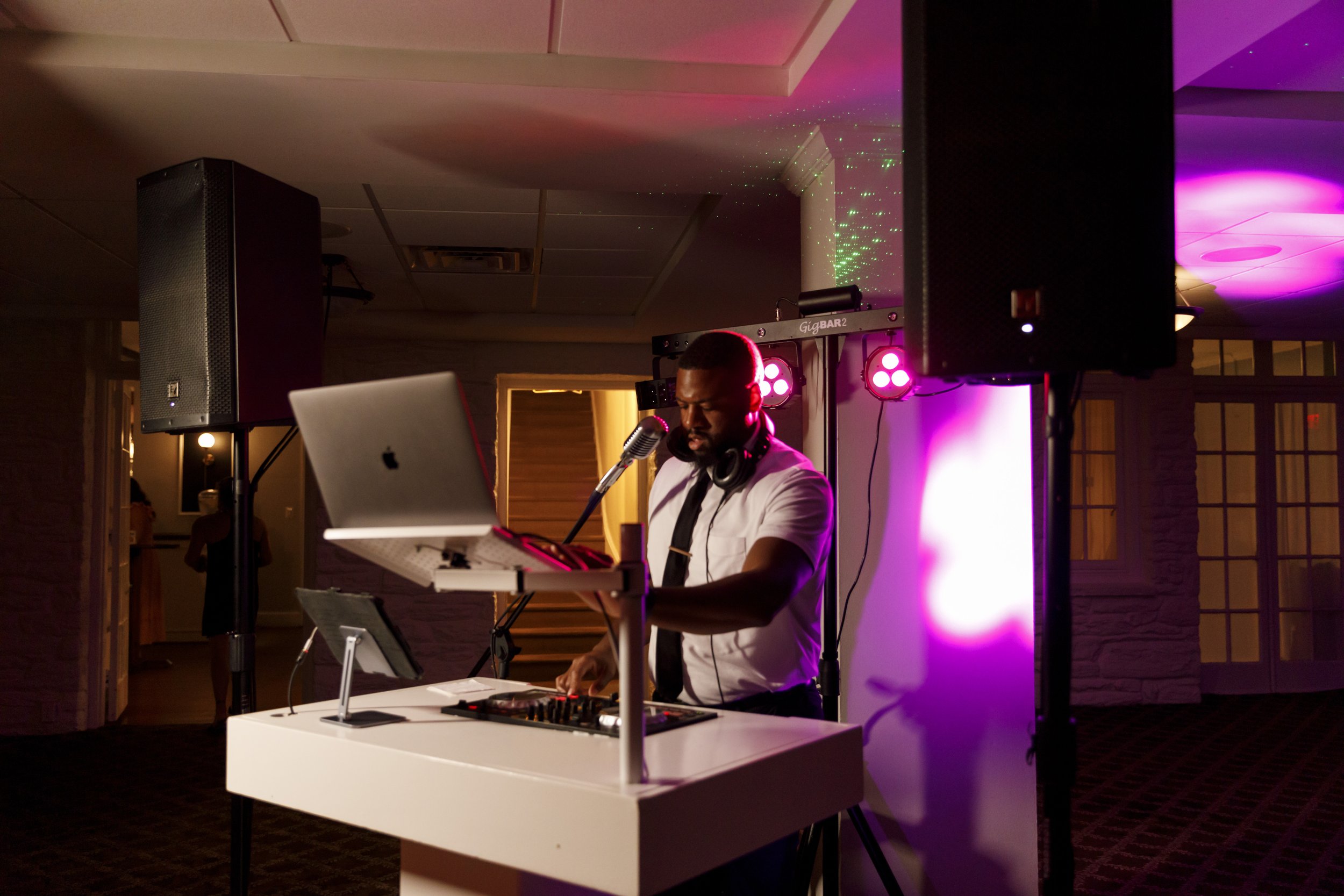 A DJ performs at a party with colorful lights, a laptop, and equipment in a decorated indoor venue.