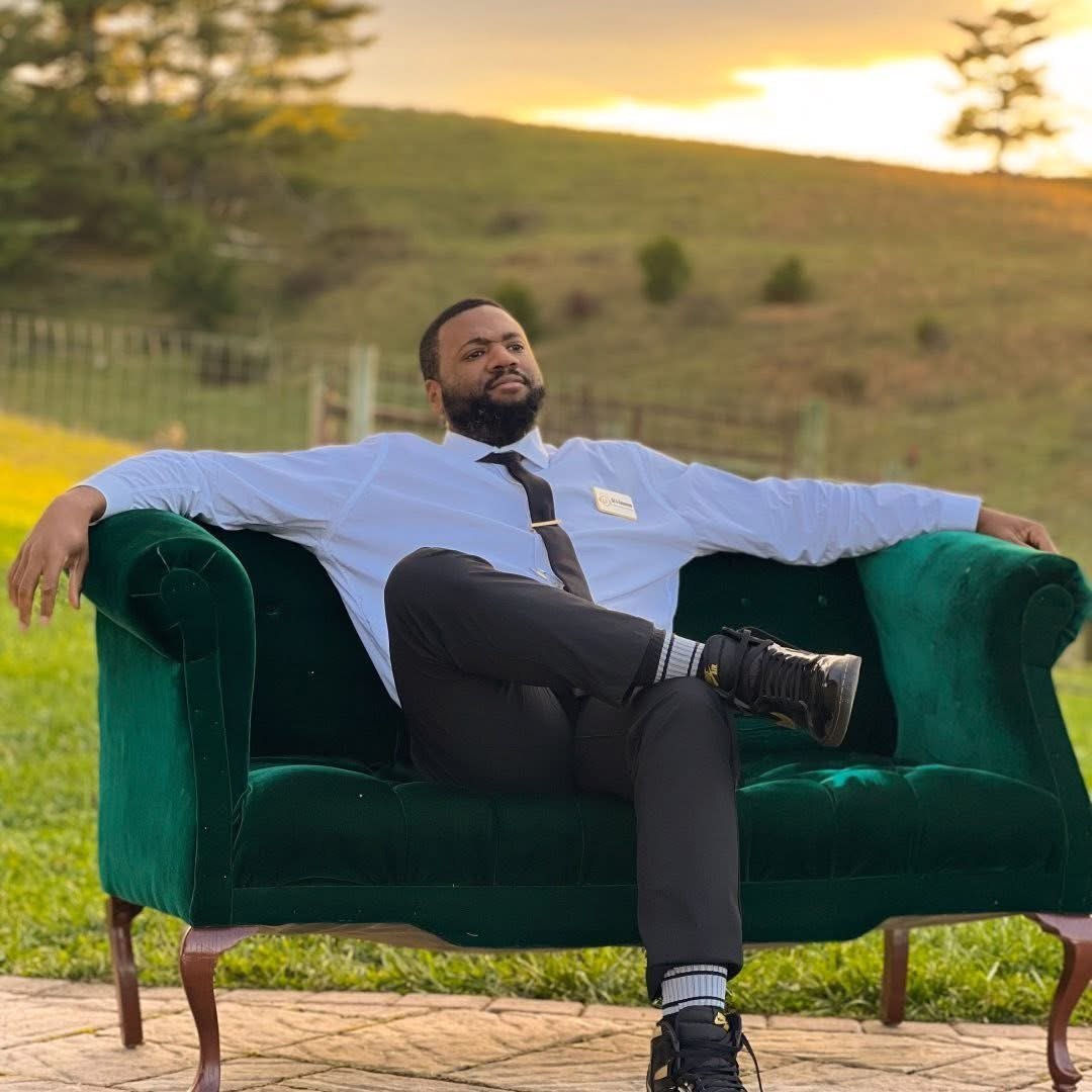 A man in formal attire with a blue shirt, black pants, and sneakers, sitting relaxed on a green velvet sofa outdoors during sunset, with a scenic grassy hill and trees in the background.
