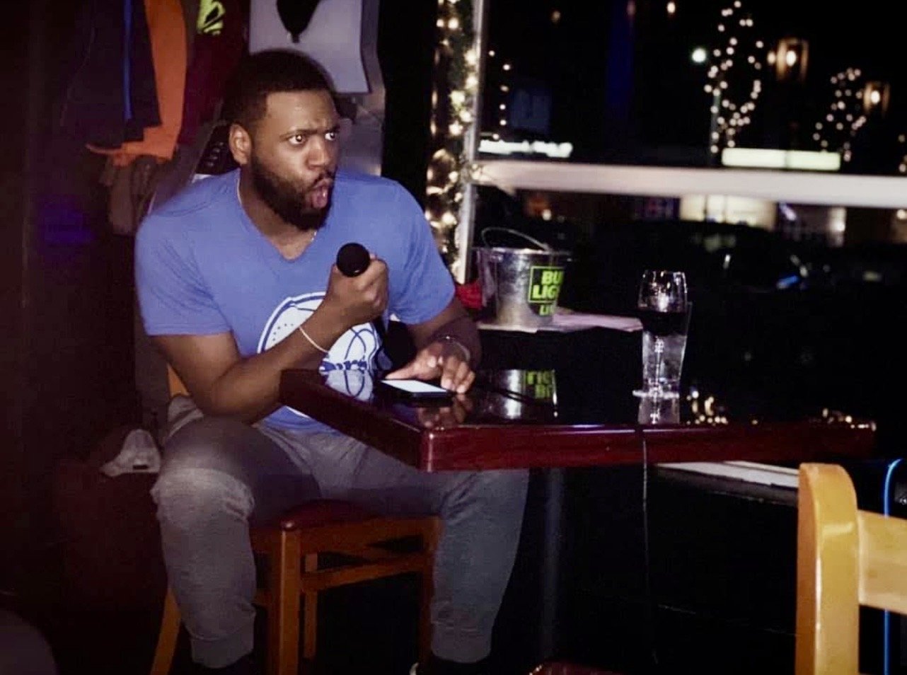 A man sitting at a table, holding a microphone, with a surprised or intense expression. There is a glass of water, a bucket with a Bud Light logo, and some other items on the table. The background shows a dark venue with string lights outside a windo