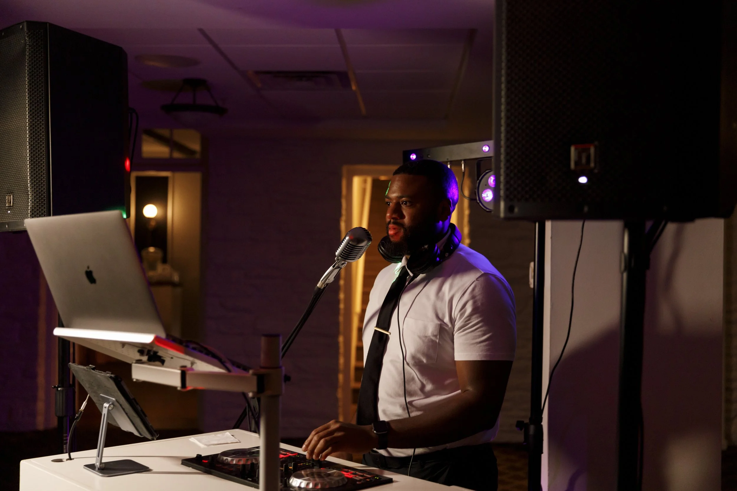 A DJ performing at a dimly lit event, wearing a white shirt, black tie, and headphones, with DJ equipment, a laptop, and speakers in the background.
