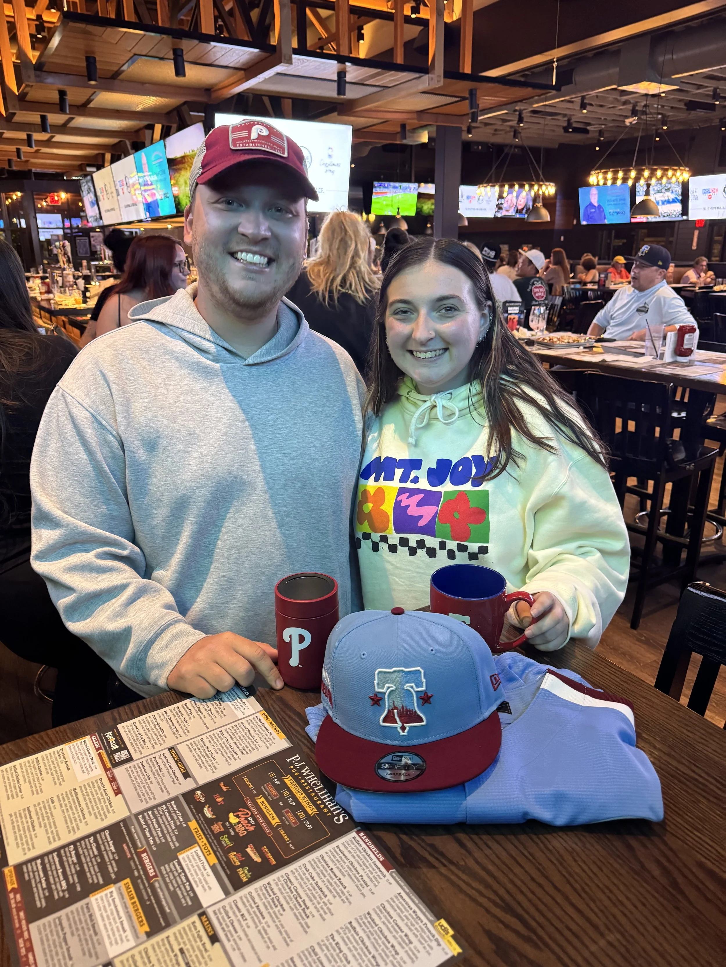 A smiling man and woman sit at a restaurant table with a Texas Rangers baseball cap, a Phillies mug, and a red travel mug on the table. The woman wears a colorful hoodie. The background shows a busy sports bar with multiple TV screens.