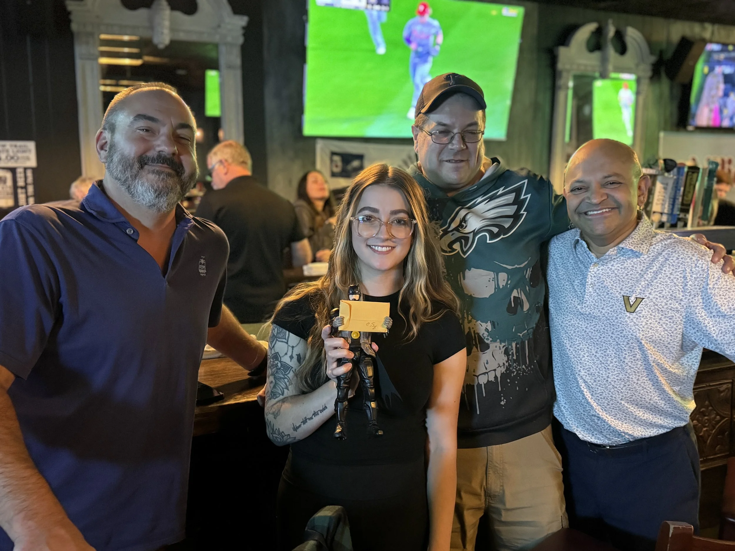 Four people are in a bar with a TV showing a football game in the background. They are smiling, with the woman holding a small hand-held game console or device.
