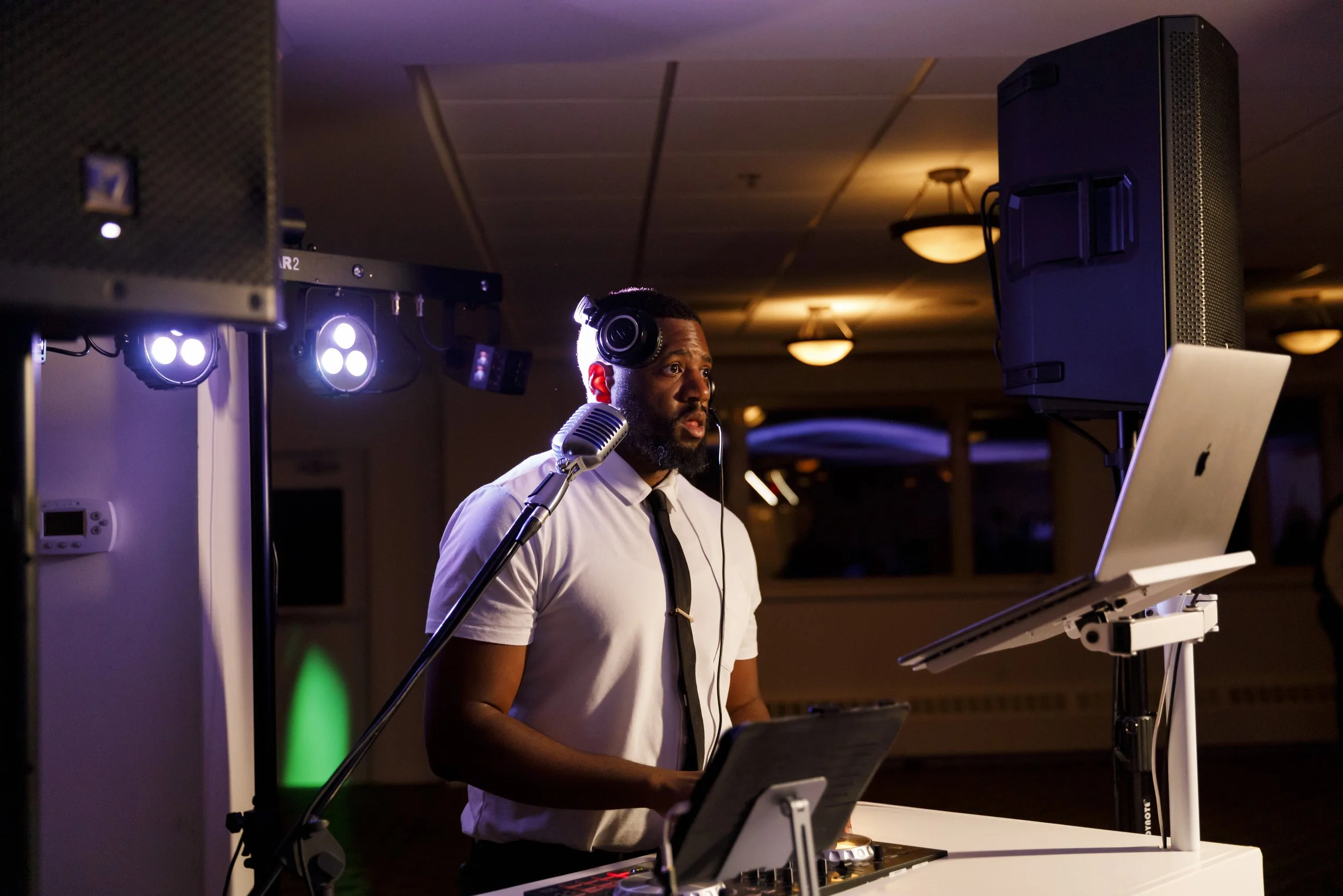 A DJ wearing headphones and a white shirt with a black tie stands behind a DJ booth with a laptop, mixer, and microphone, in a dimly lit event space with colorful lighting.