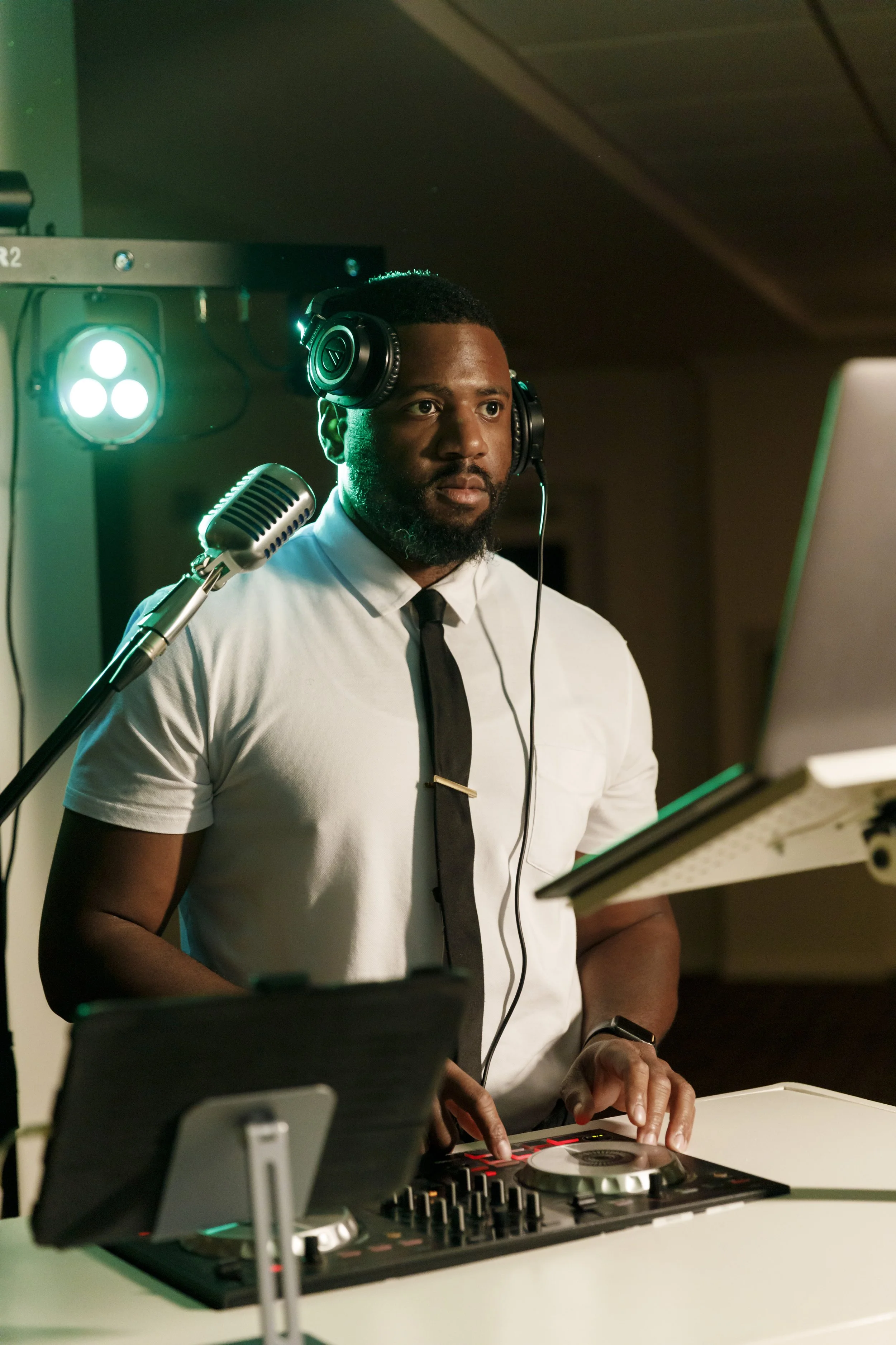 A DJ wearing headphones, a white shirt, and a black tie, standing at a DJ controller with a laptop, in a dimly lit room with green stage lights.