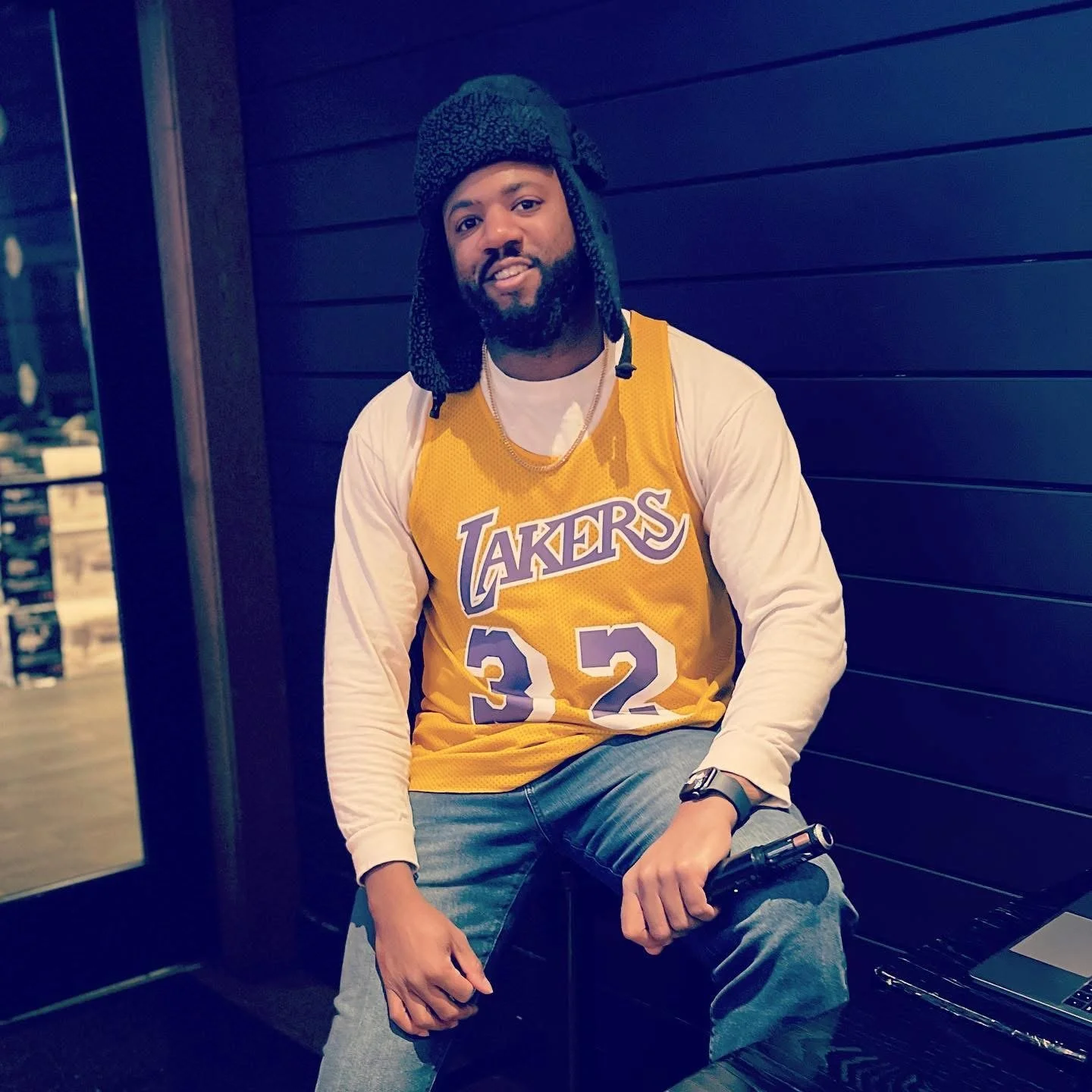 A man with a beard wearing a yellow Los Angeles Lakers basketball jersey with the number 32, a white long-sleeve shirt underneath, blue jeans, a black watch, and a black head covering, sitting with a microphone in his hand, in front of a dark wooden wall.