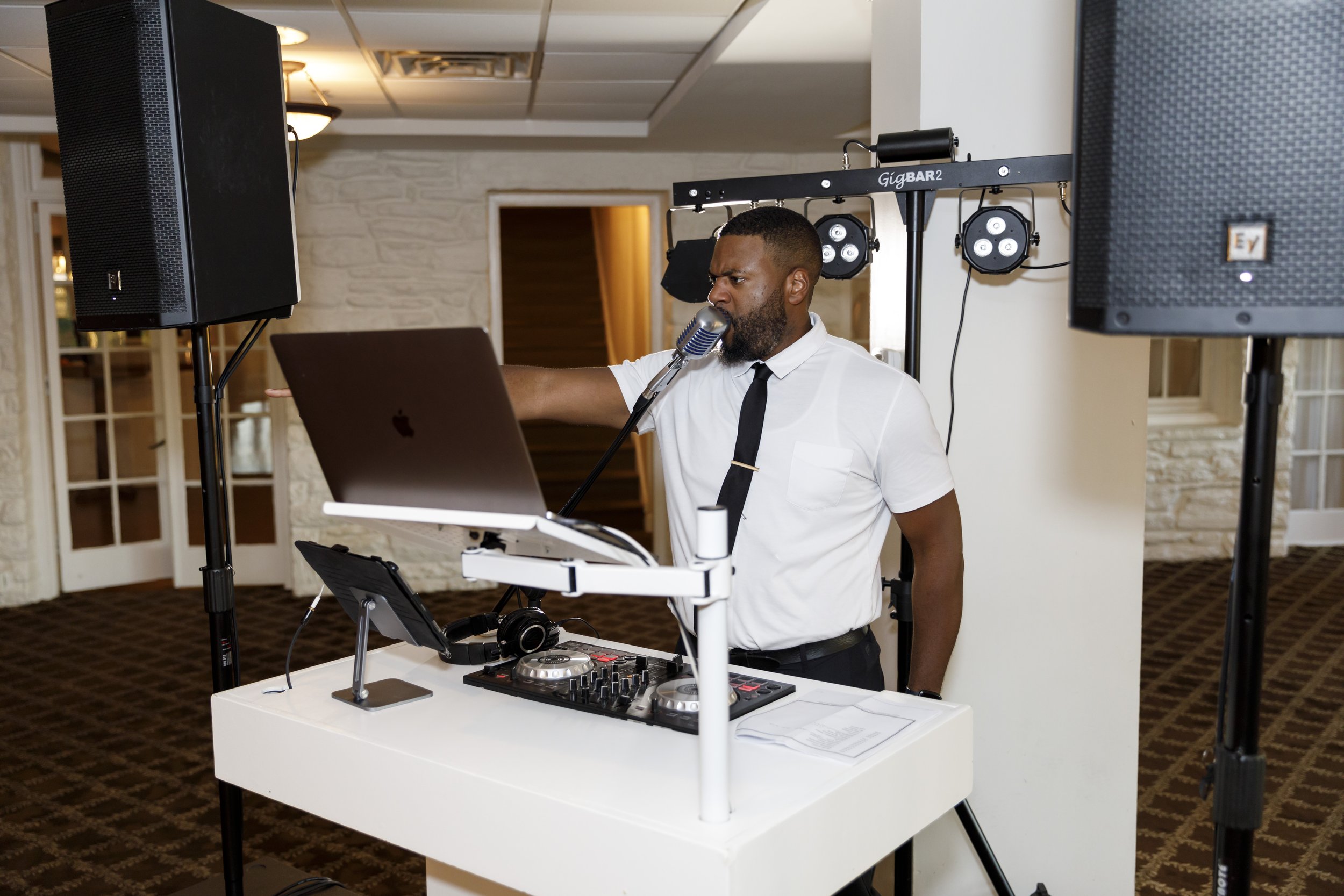 A male DJ in a white shirt and black tie performs at an event, standing behind a white DJ booth equipped with a laptop, a DJ controller, and headphones, with speakers and stage lighting in the background.