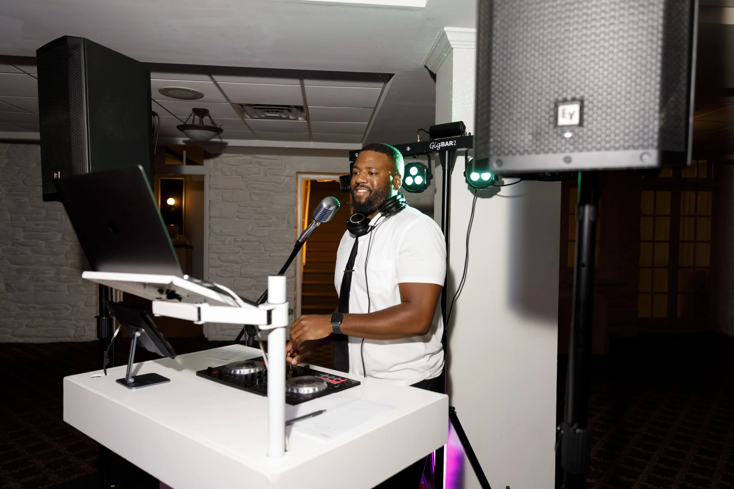 A man with a beard and headphones around his neck DJing at an event, smiling, standing behind a white DJ booth with a laptop and DJ equipment, with professional lighting and large speakers in the background.
