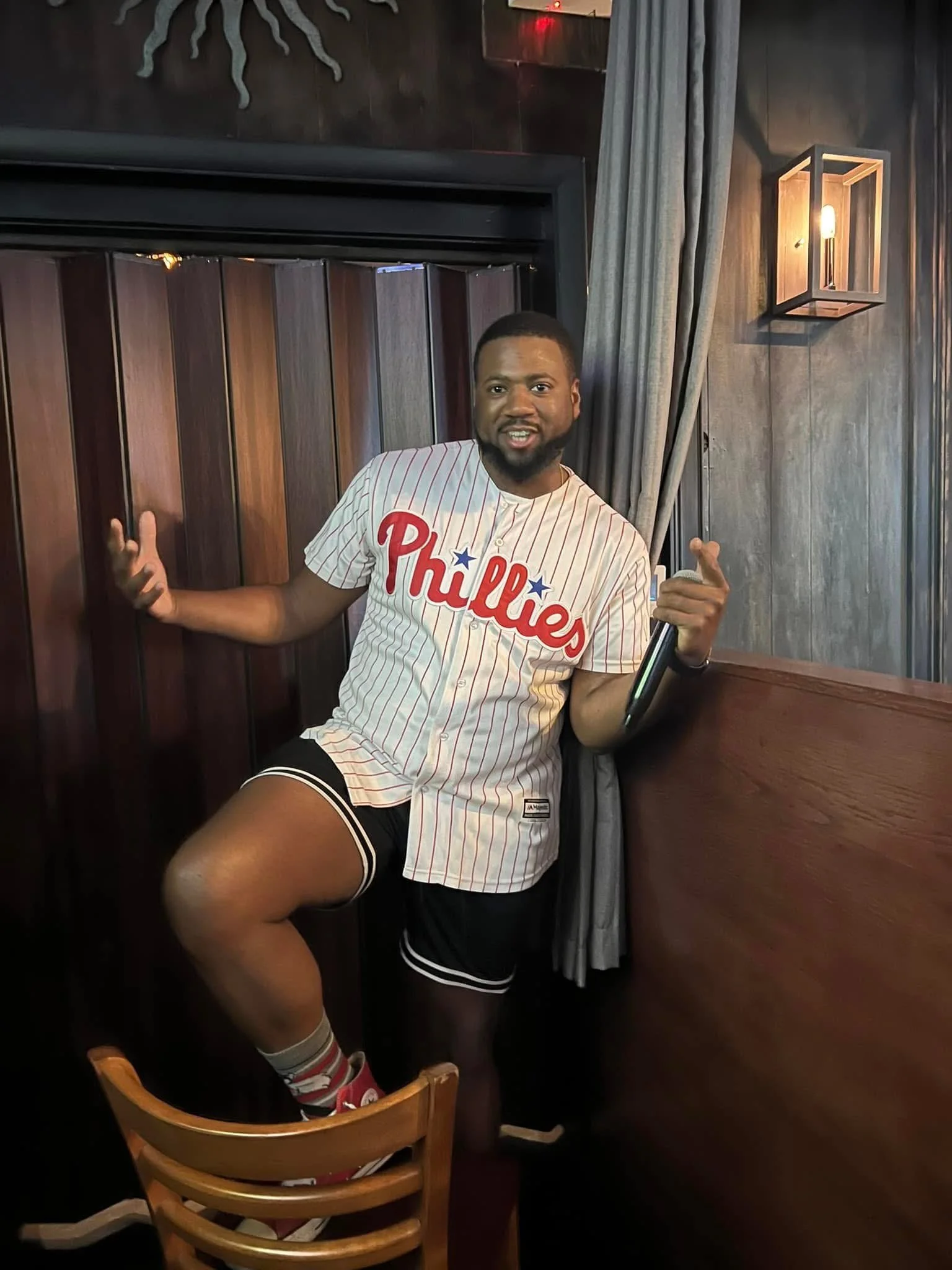 A man dressed in a Philadelphia Phillies baseball jersey, shorts, and Phillies socks, standing in a cozy, dimly lit room with wooden walls, posing with one foot on a chair and gesturing with his hands.