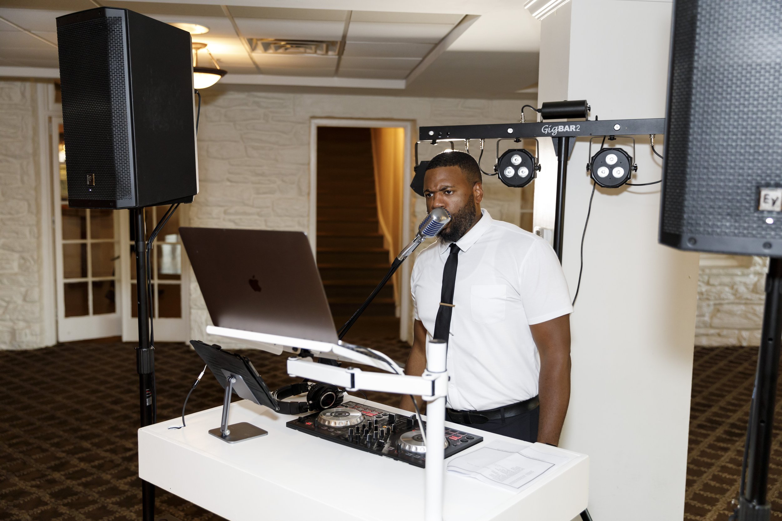 A DJ in a white shirt and black tie performs at an indoor event, surrounded by speakers, a laptop, and DJ equipment.