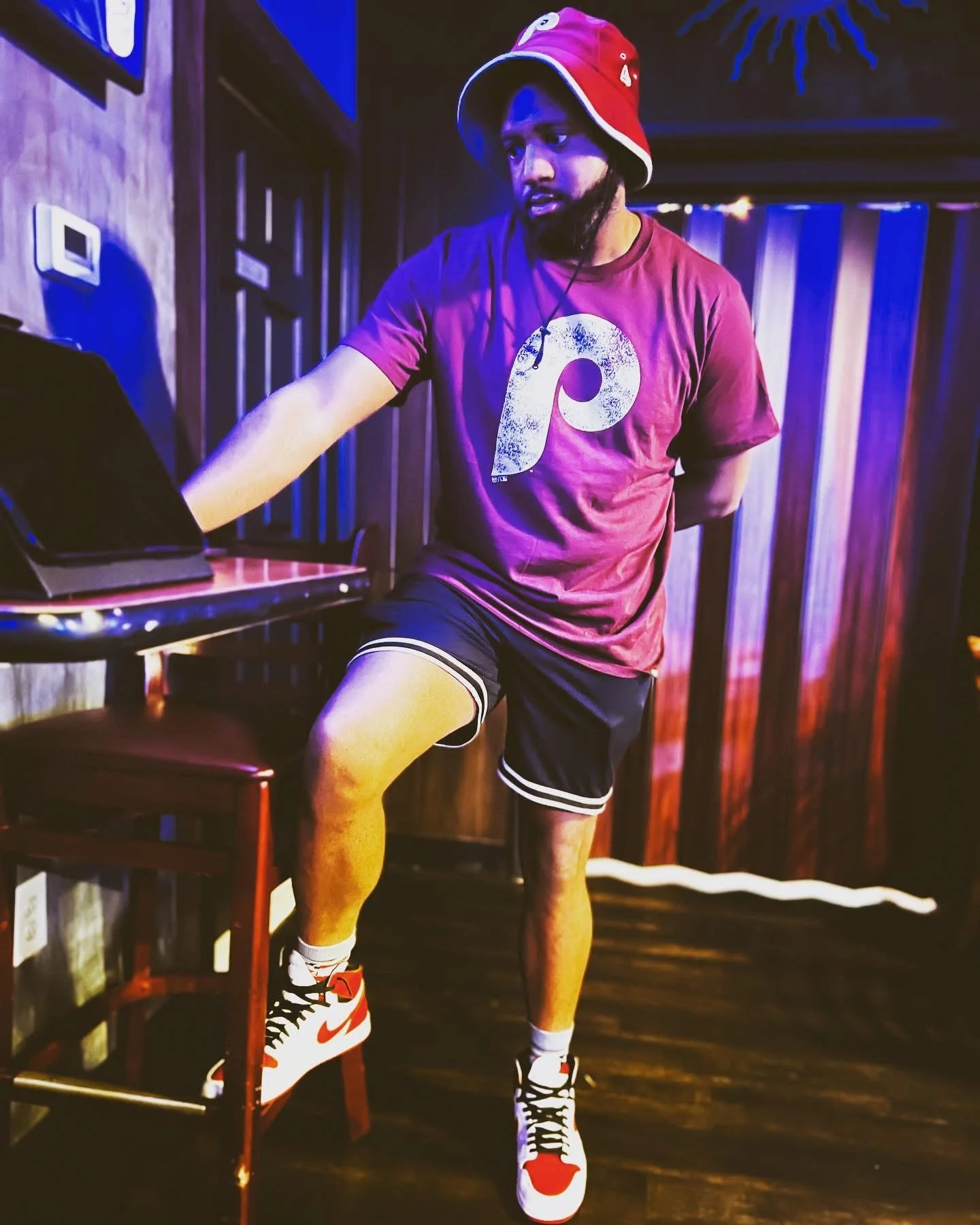 A man in a maroon T-shirt, black shorts, and red, white, and black Nike sneakers is standing with one foot on a chair, in a dimly lit room with purple and blue lighting. The man is wearing a red bucket hat and has a beard. He appears to be DJing or w