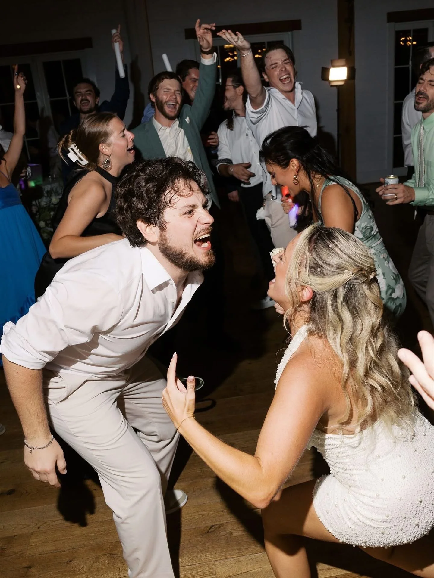 People dancing and singing at a lively party or wedding reception, with some holding drinks and decorations, expressing joy and excitement.