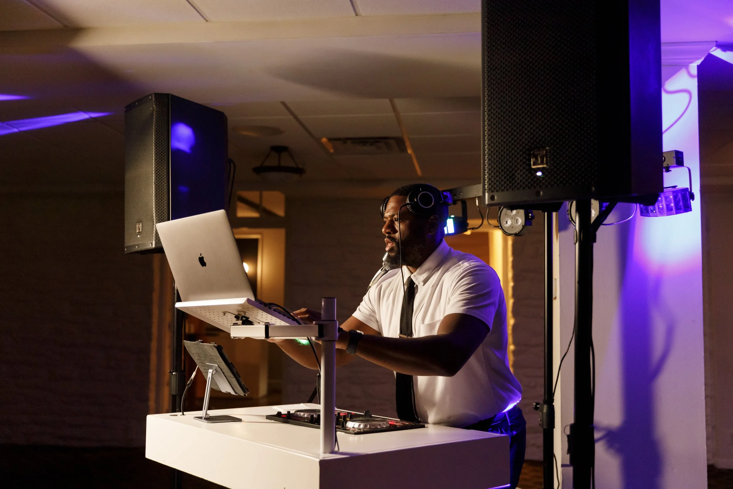 A DJ in a white shirt and black tie wearing headphones, operating music equipment with a laptop in a dimly lit indoor venue with large speakers and purple lighting.